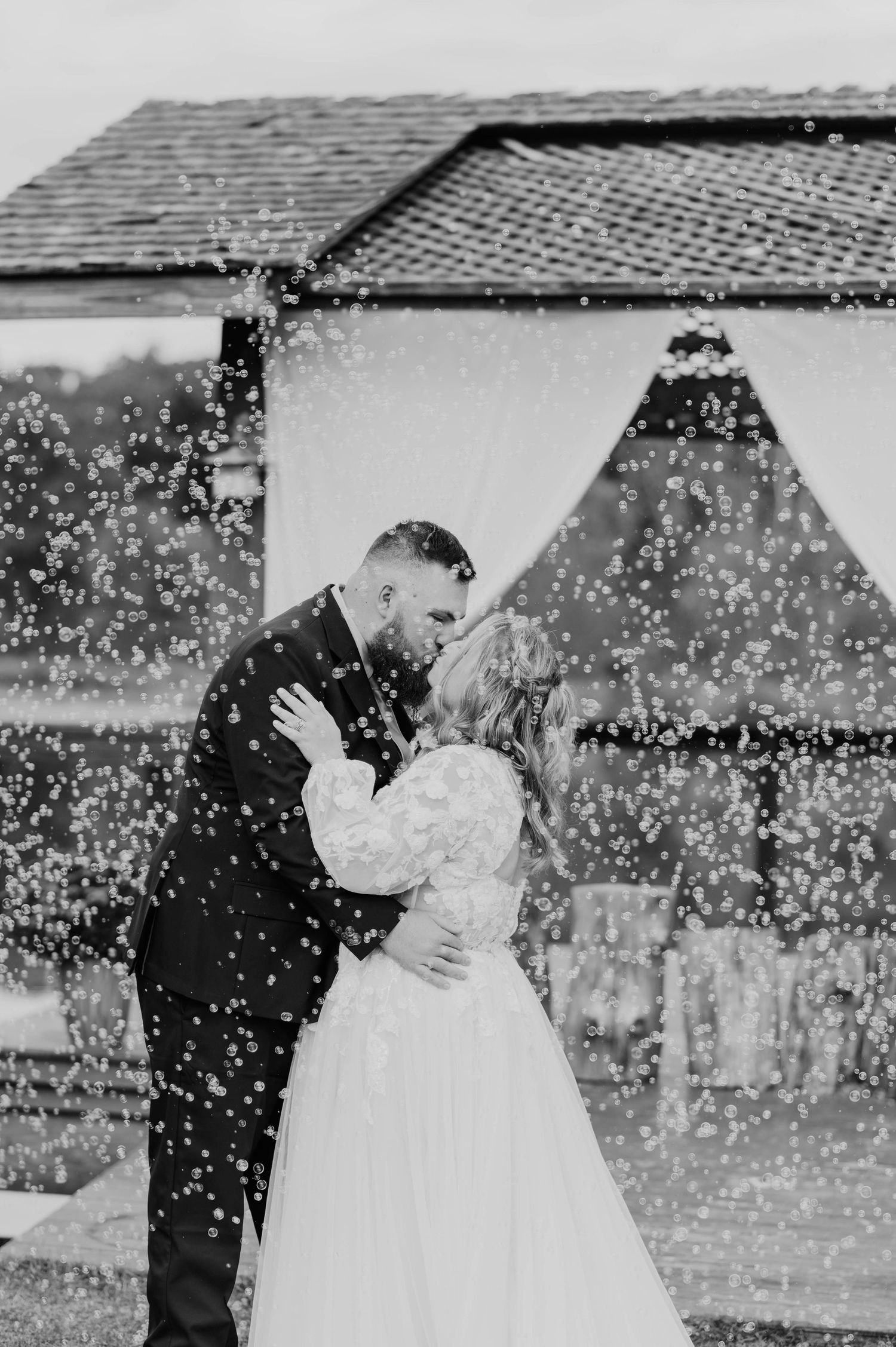 Black and white photo of newlyweds sharing first kiss surrounded by bubbles at outdoor ceremony.