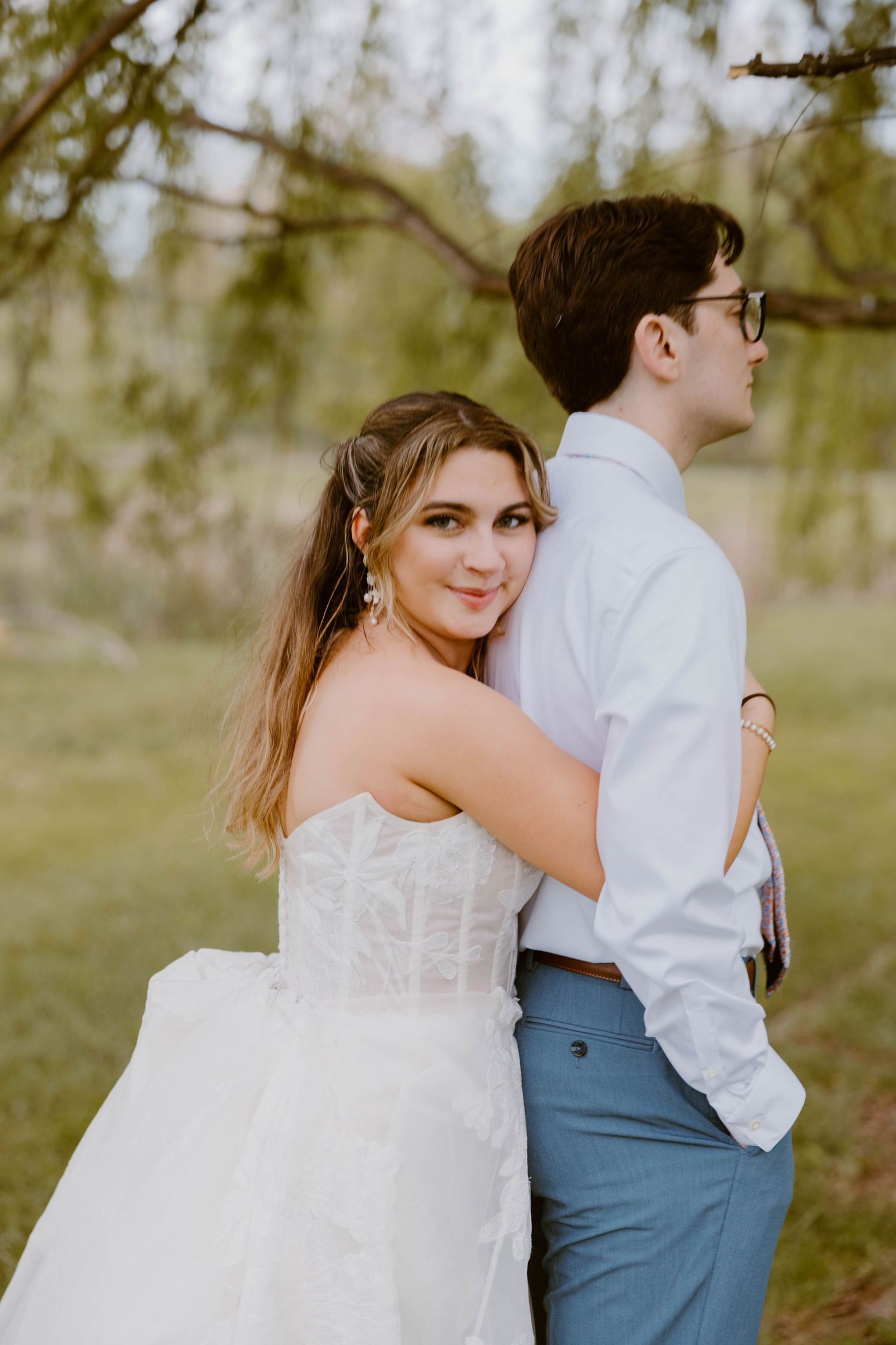 Bride in strapless white ball gown embraces groom from behind outdoors, smiling at camera in a lush green setting.
