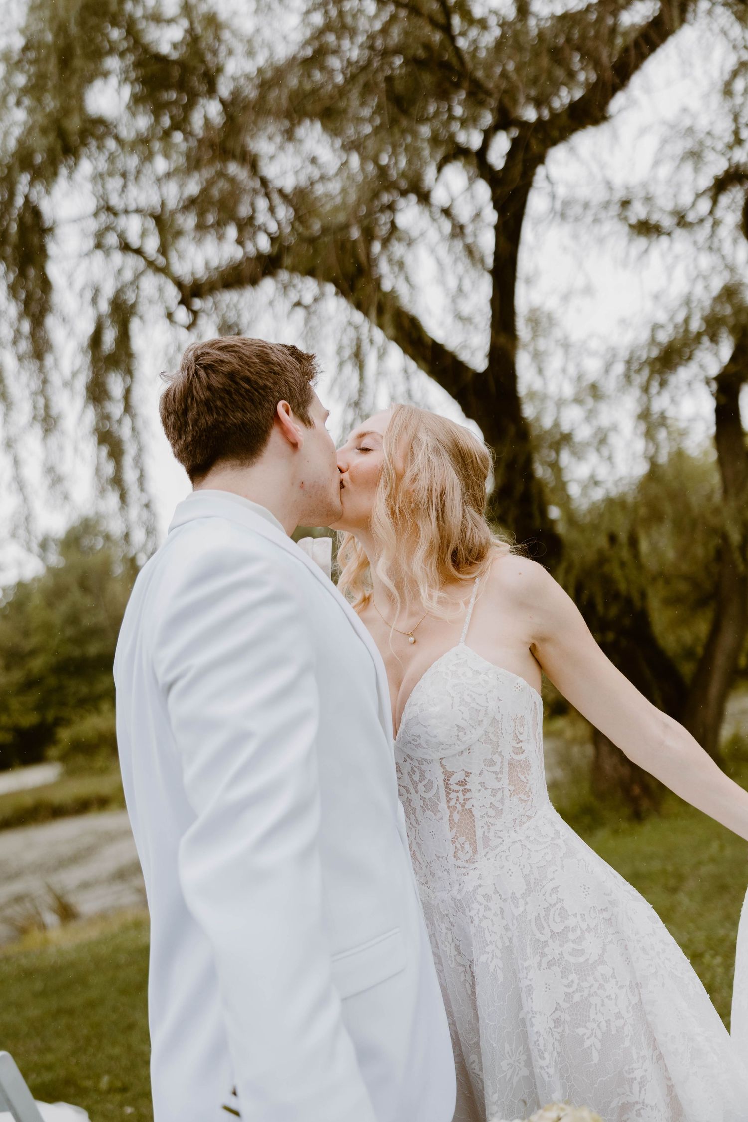 Bride and groom share a romantic kiss outdoors under a willow tree, bride in spaghetti strap lace gown, groom in white suit.
