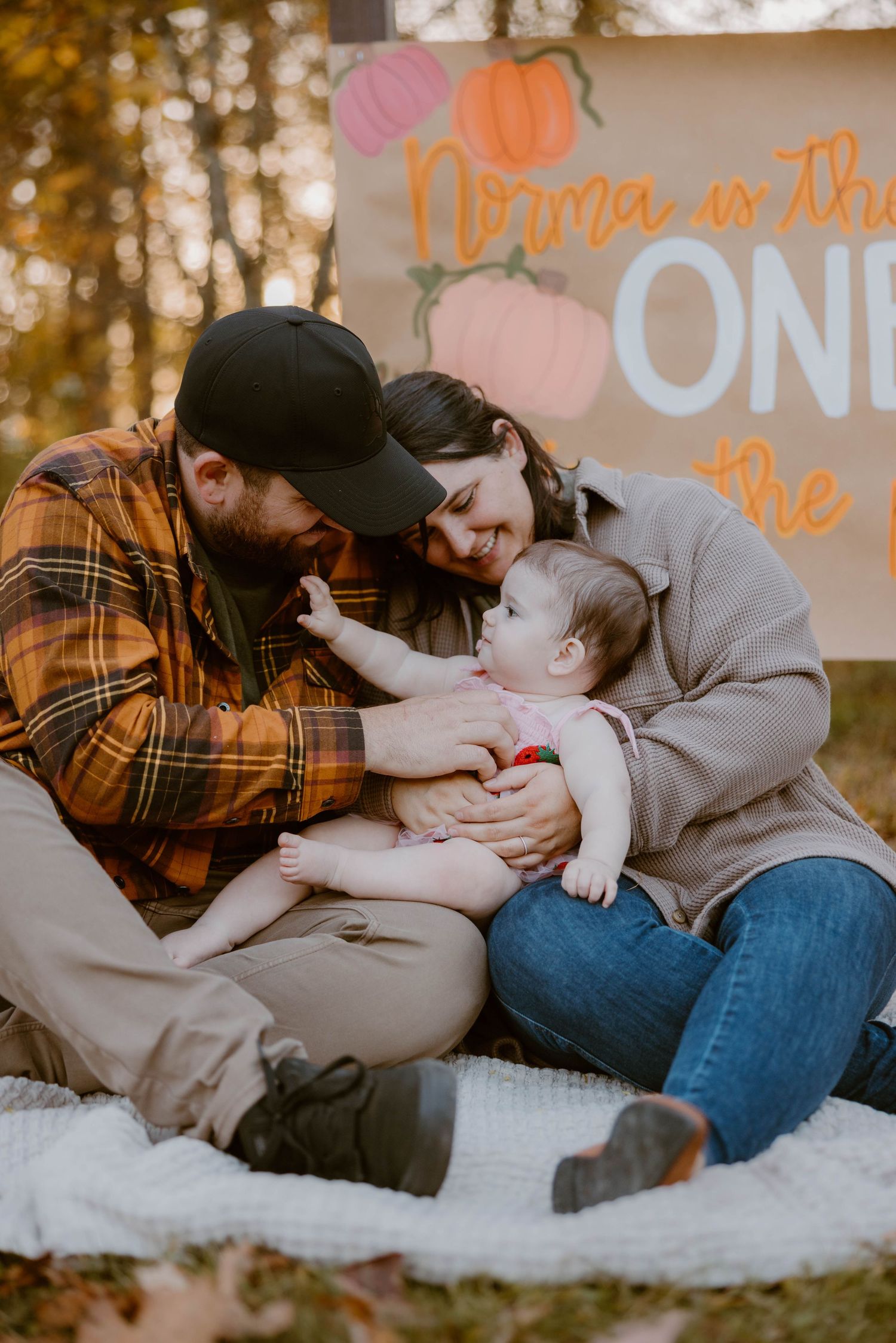 Happy family sits together on a blanket with a pumpkin-themed birthday sign in autumn woods.