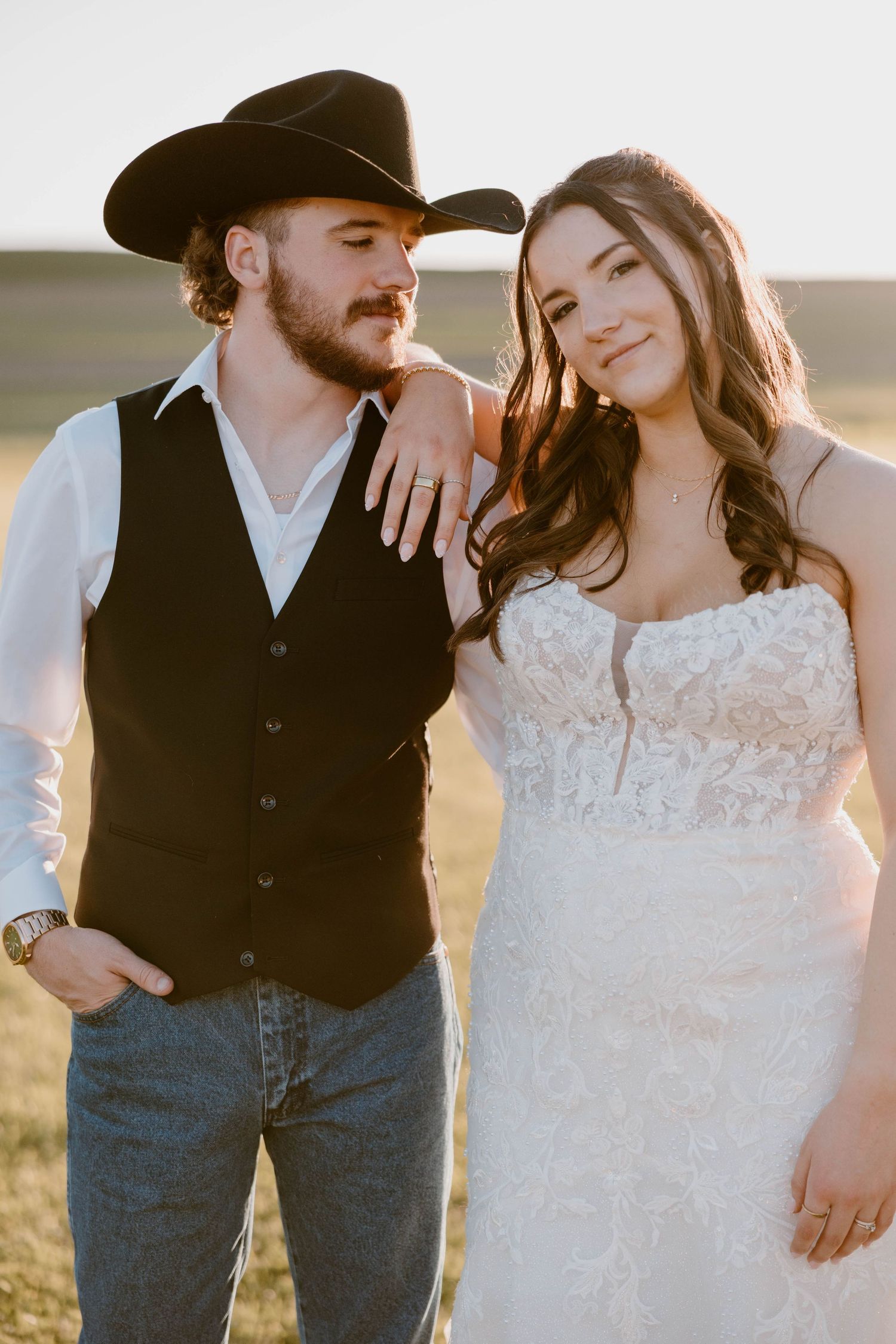 Bride and groom pose outdoors at golden hour, groom wearing cowboy hat and vest, bride in strapless lace wedding gown.