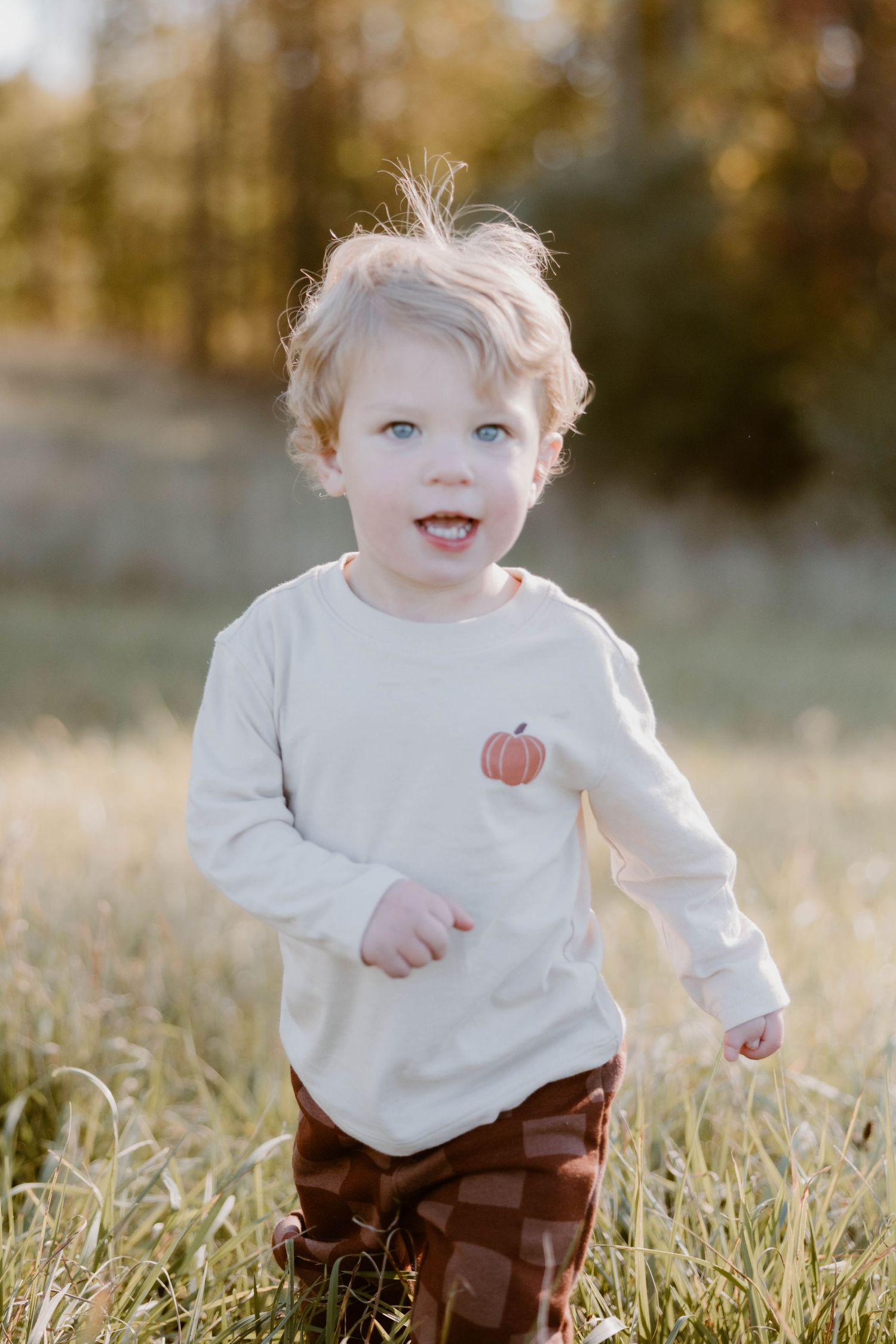 Adorable curly-haired toddler in a pumpkin shirt runs through a sunlit golden grass field.