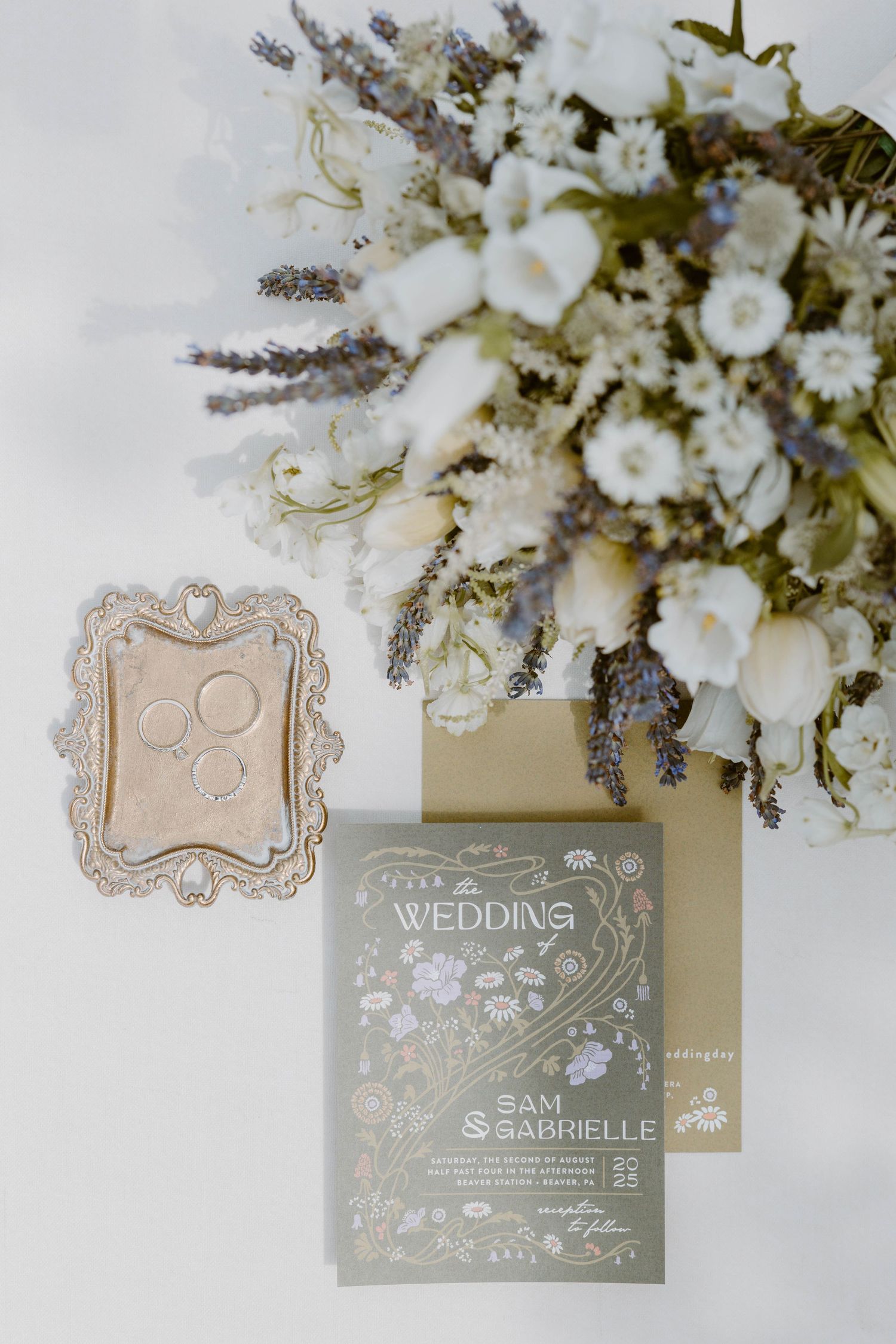 Elegant wedding flat lay with white lavender bouquet, gold rings, and illustrated Sam and Gabrielle invitation.
