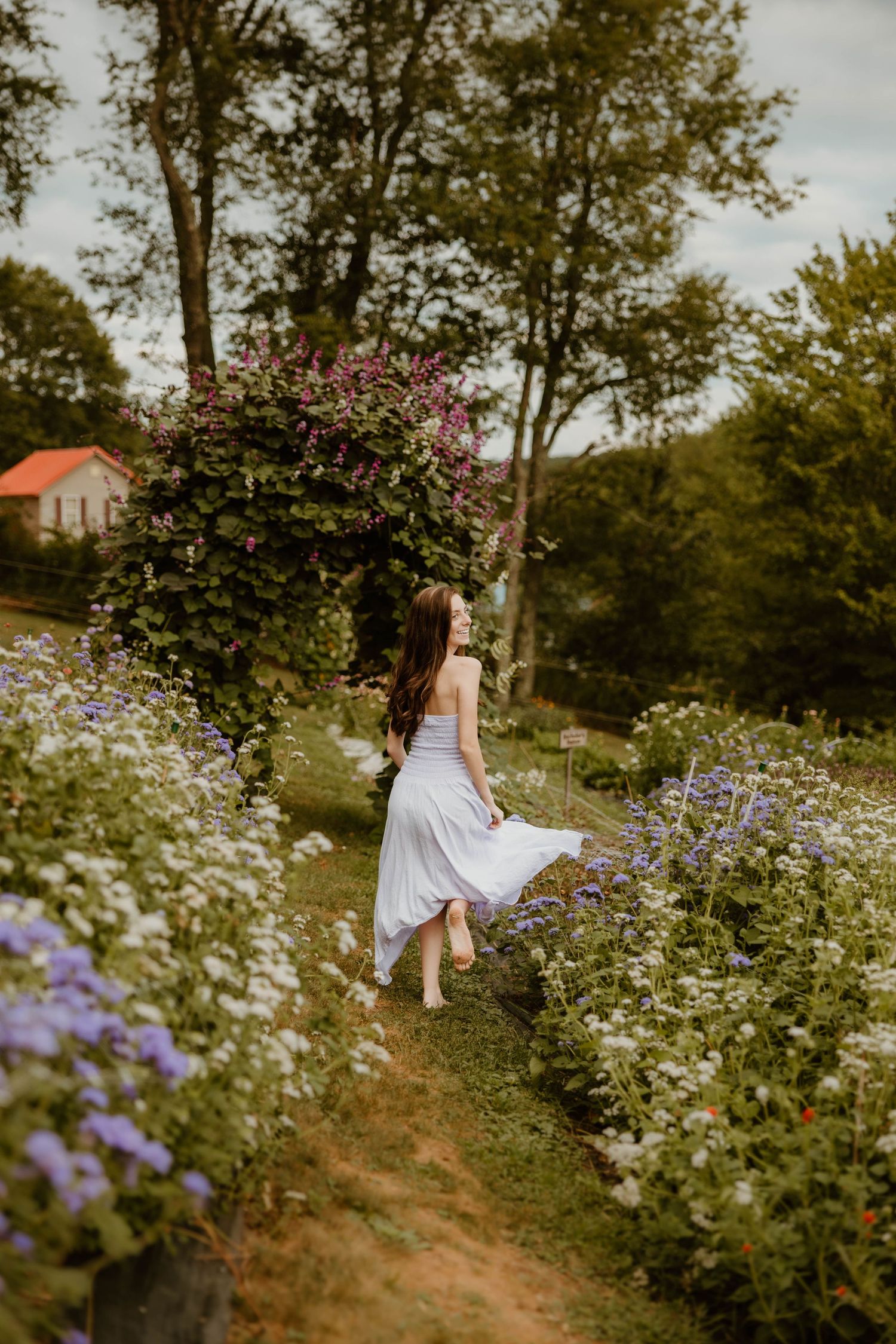 Young woman in white dress walking through a lush garden path surrounded by colorful wildflowers and blooming shrubs.