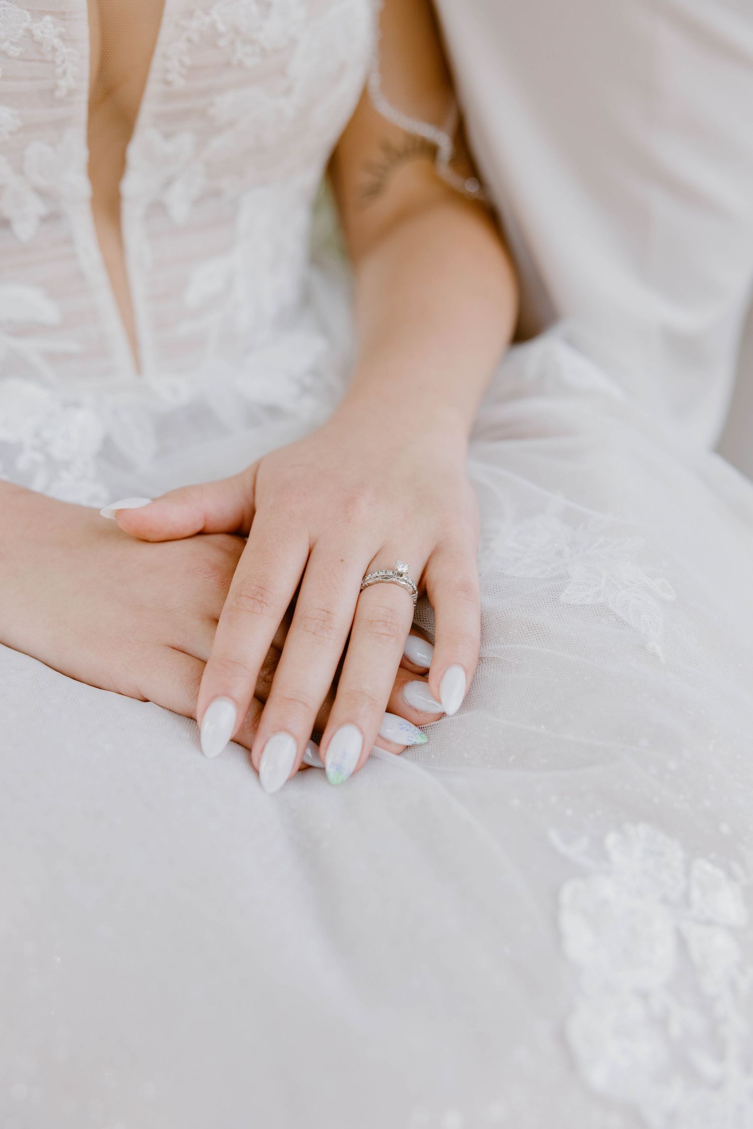 Close-up of bride's hands folded on white lace wedding dress, wearing a delicate diamond engagement ring.