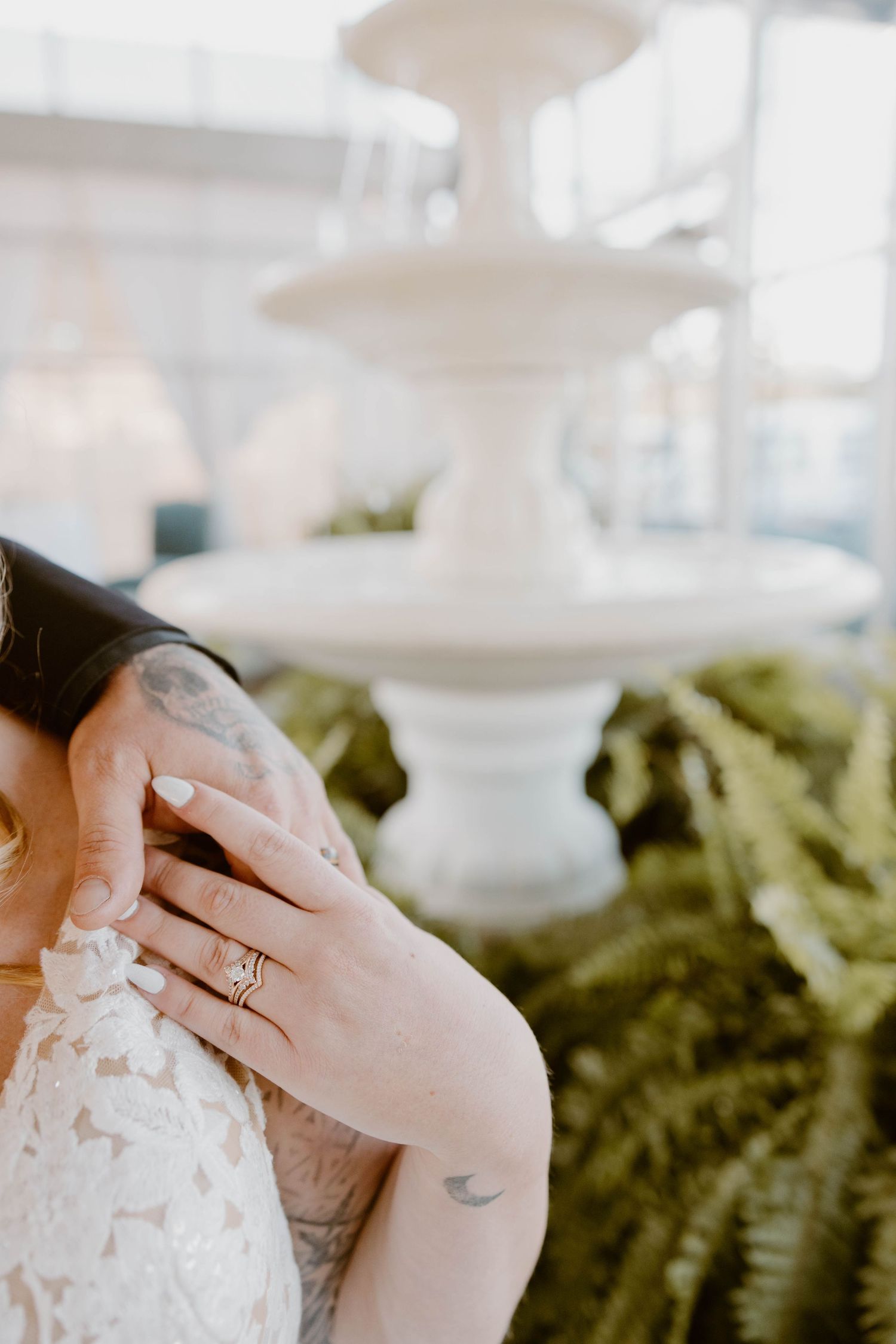 Close-up of bride wearing a diamond engagement ring, grooms tattooed hand gently cradling her face near a fountain at the Atrium in Butler,