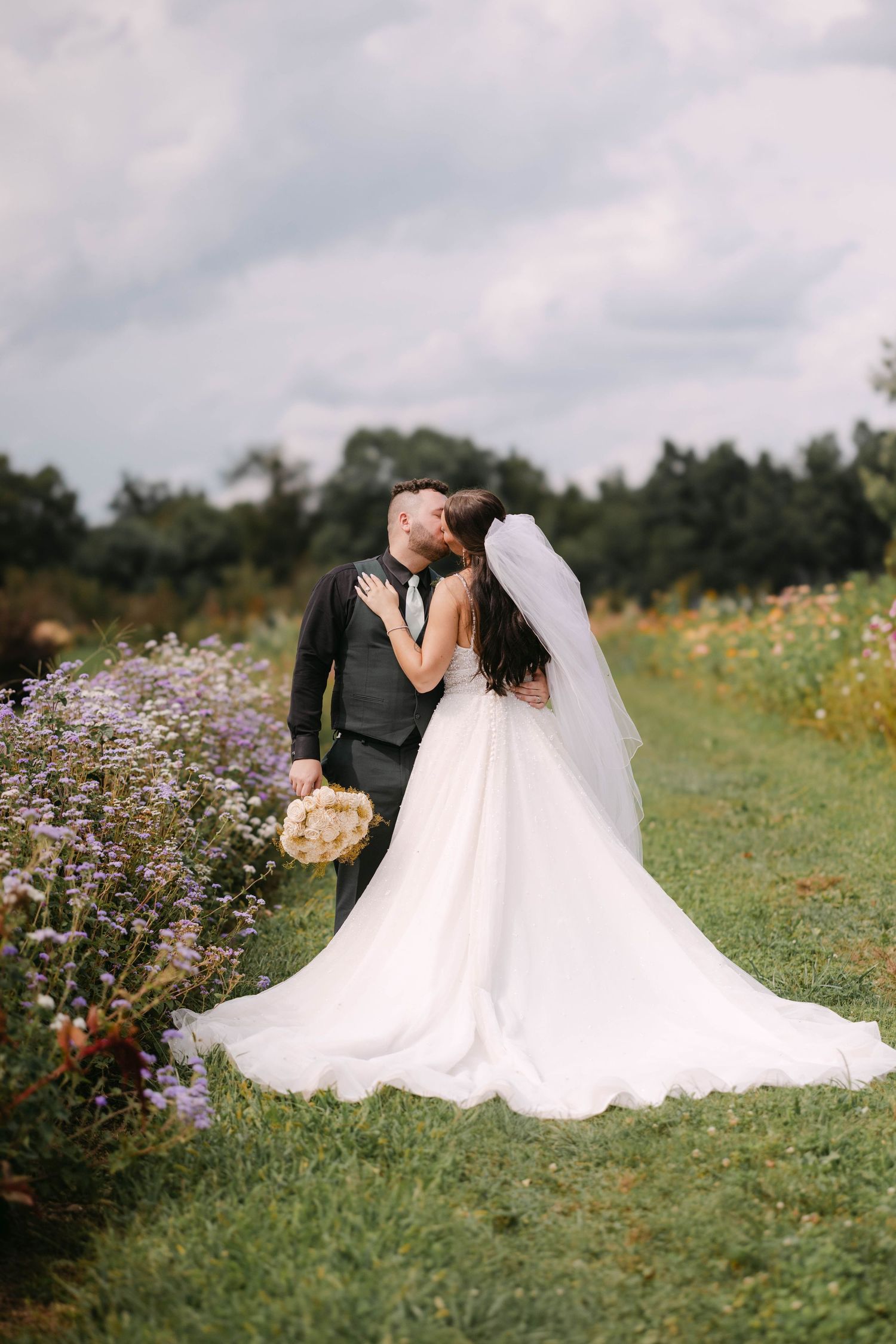 Bride in a flowing white gown and veil kisses her groom in a vest and tie while standing in a flower field under a cloudy sky.