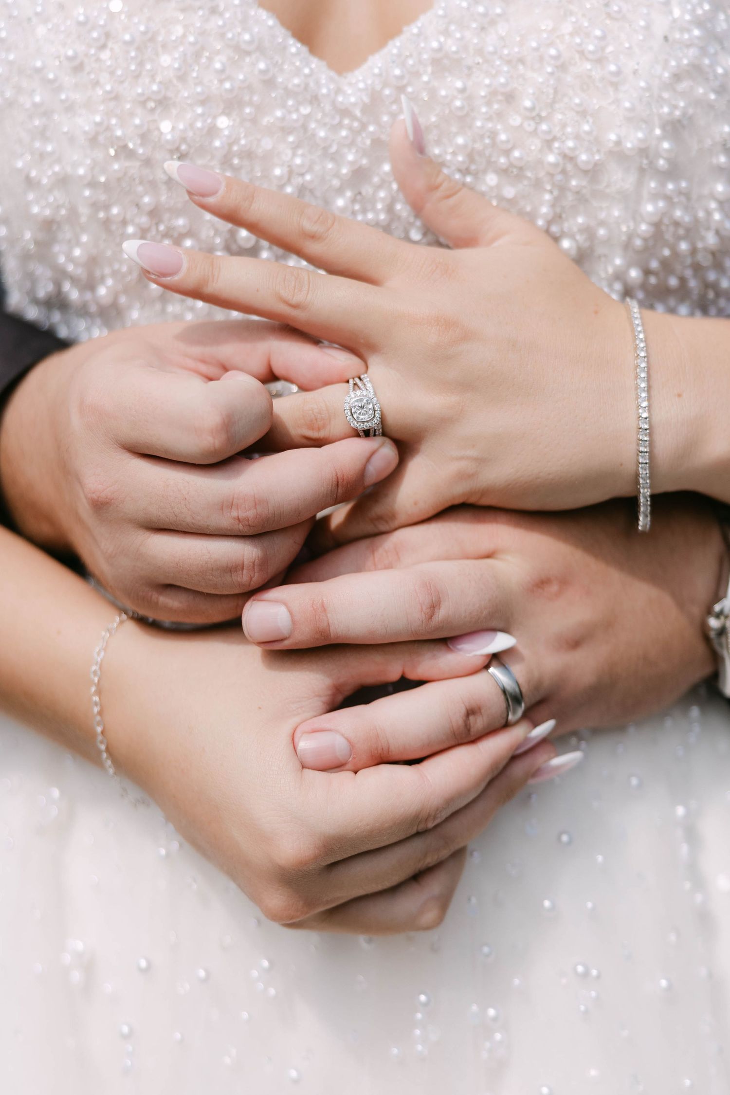 Close-up of a bride's hands wearing a diamond engagement ring and bracelet, clasped with her partner's hands against a beaded wedding gown.