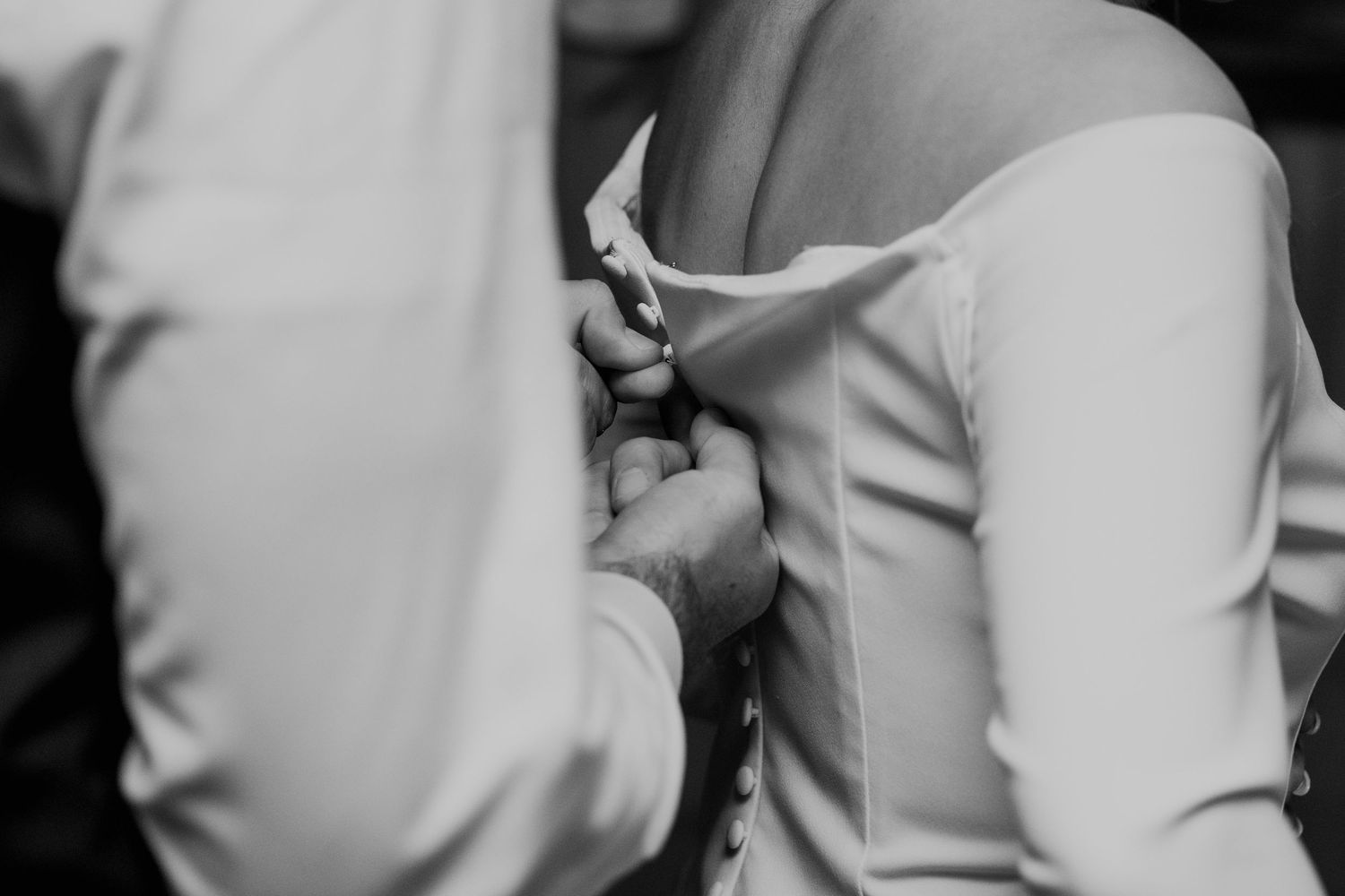 Close-up black and white photo of someone buttoning the back of a bridal gown before the wedding ceremony.
