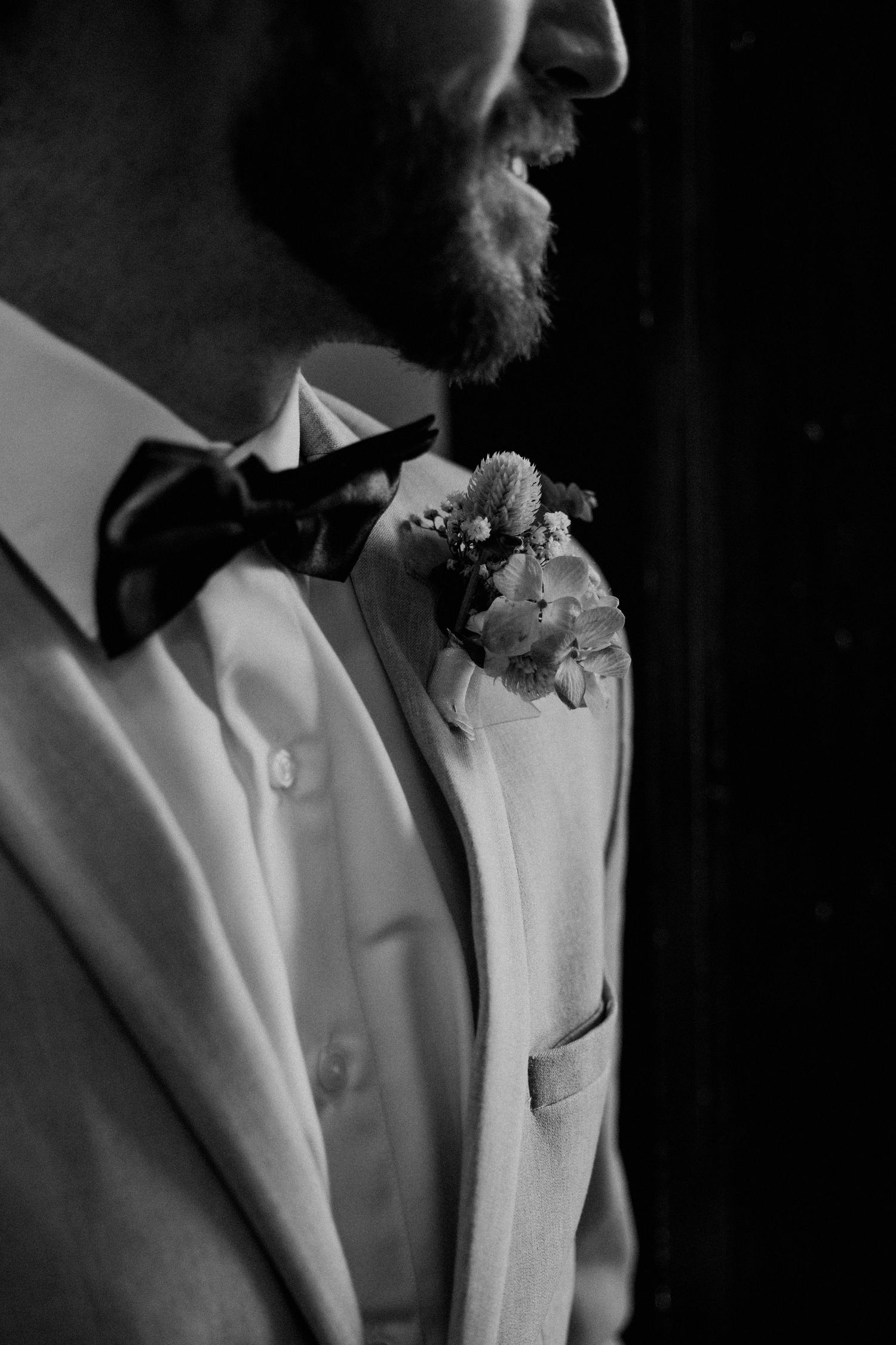 Black and white close-up of smiling groom in white suit with bow tie and floral boutonniere.