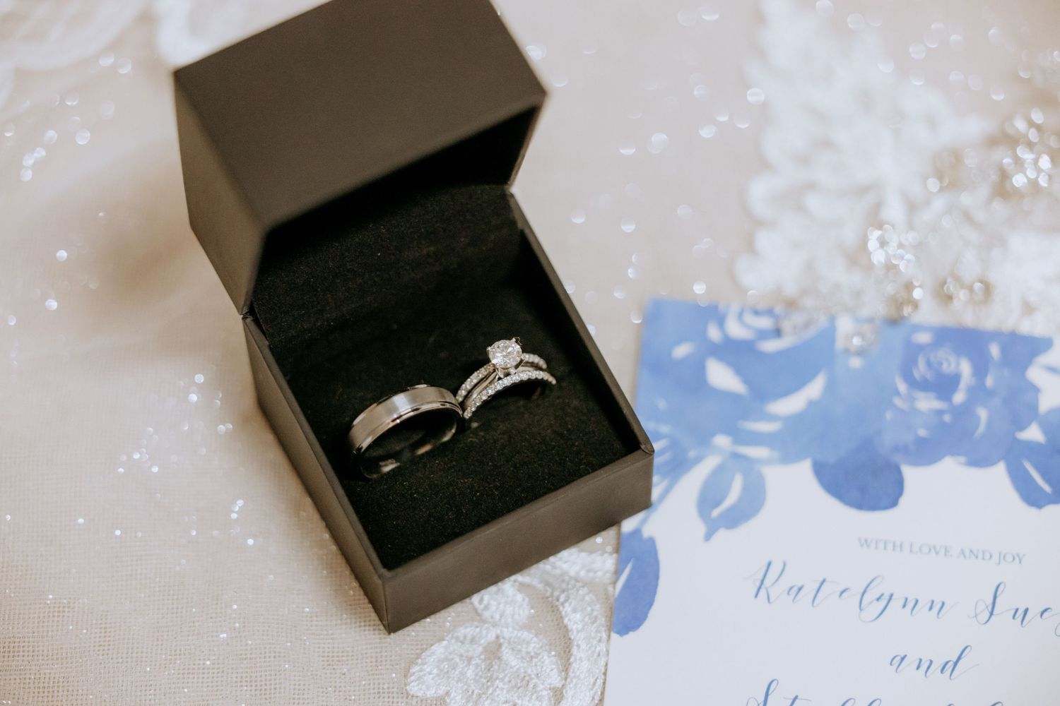 Diamond wedding ring set displayed in a black box beside a blue floral wedding invitation.