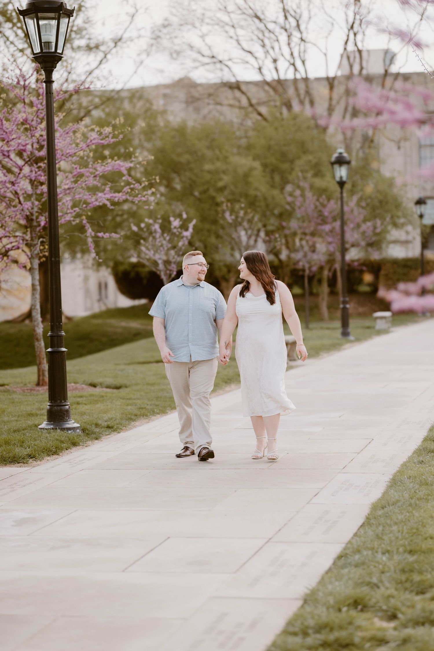 Couple holding hands walking on a scenic pathway surrounded by blooming pink trees in spring.