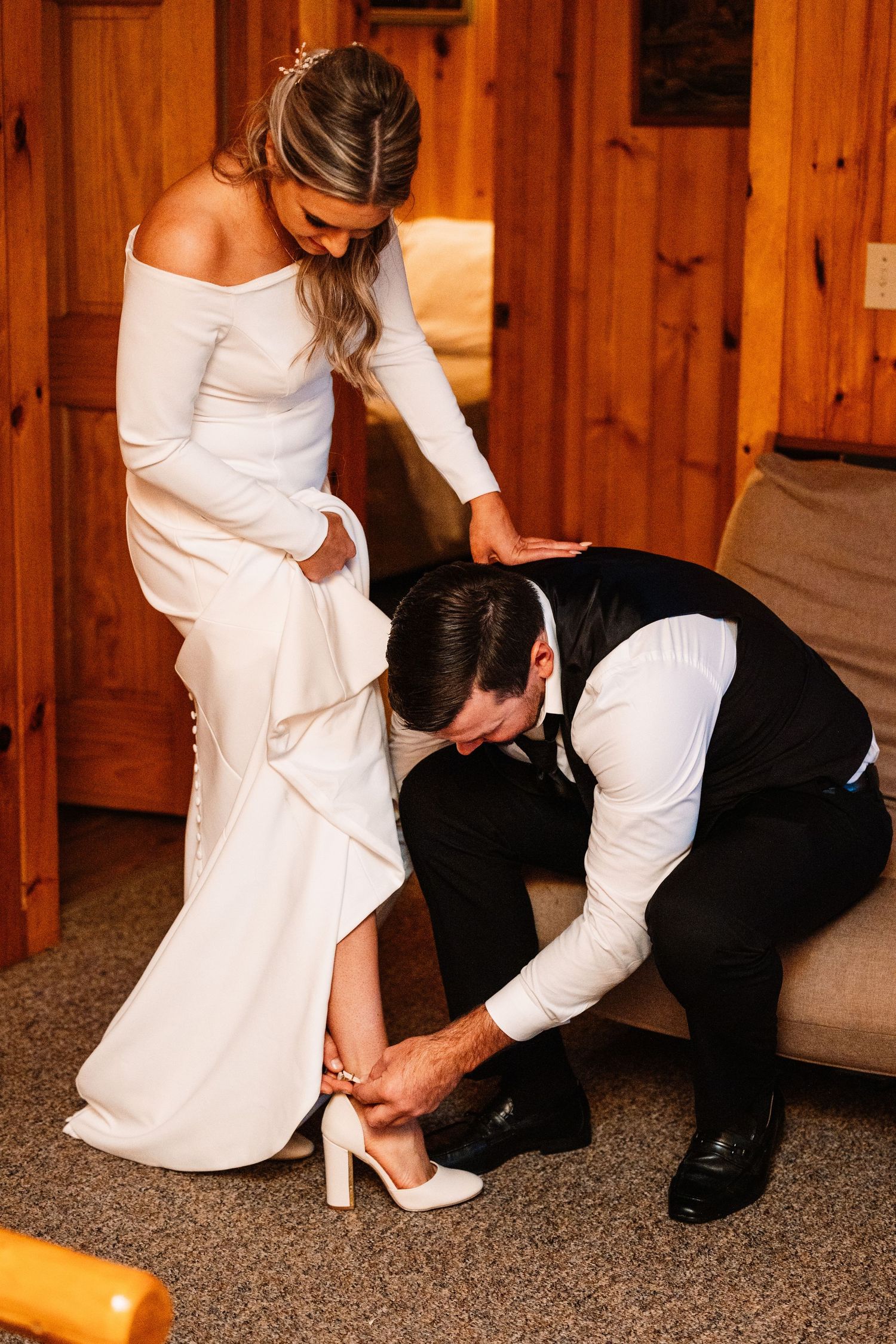 Groom kneeling to help bride put on white heeled shoes in a cozy wooden cabin.
