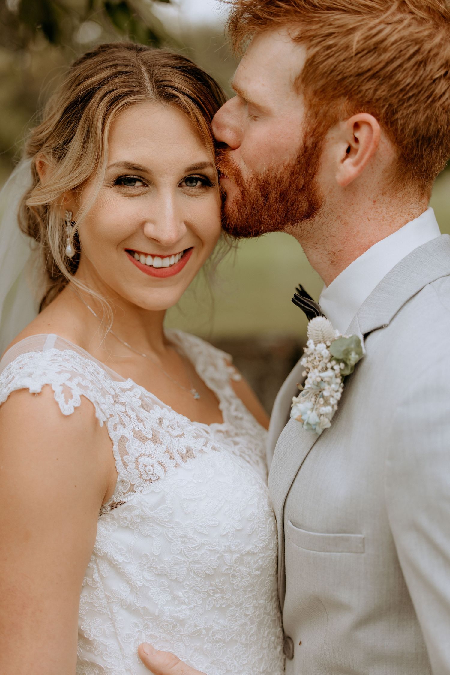 Groom with red beard tenderly kisses bride on cheek as she smiles beautifully in lace wedding gown.