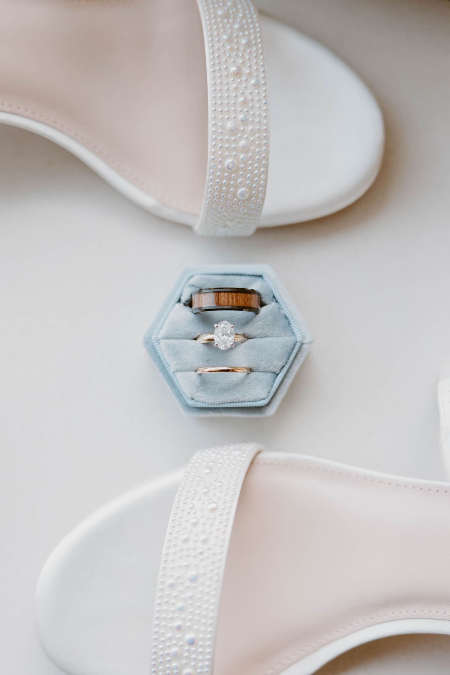 Wedding rings displayed in a light blue hexagon velvet ring box beside pearl-adorned white bridal heels.