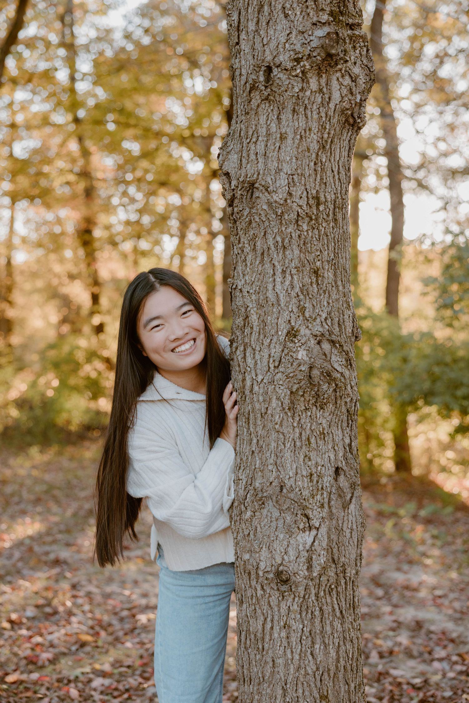Smiling young woman peeks playfully from behind a tree trunk in a sunlit autumn forest.