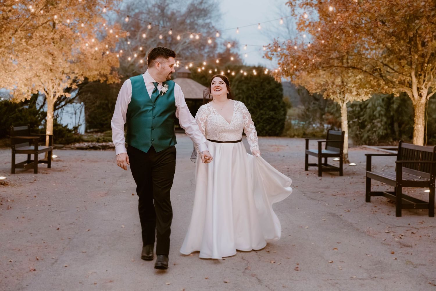 Couple walking hand-in-hand under string lights in autumn outdoor wedding venue.