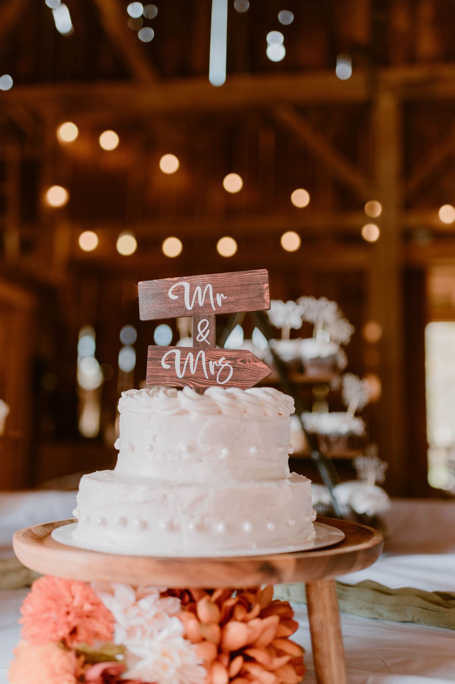 Two-tier white wedding cake with a rustic wooden Mr. and Mrs. topper inside a barn with string lights.