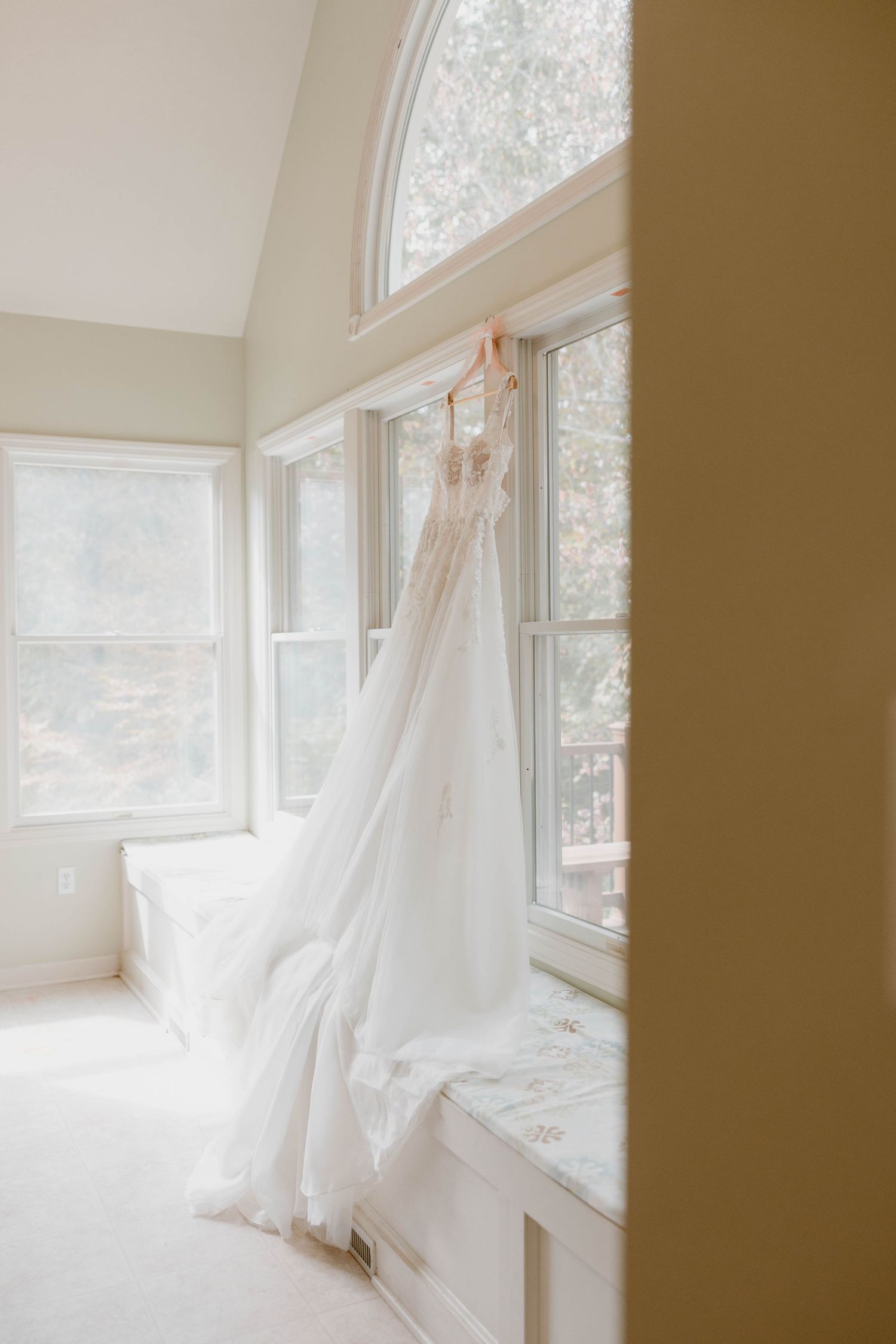 Elegant white wedding gown hanging in a sunlit window with arched glass panes.