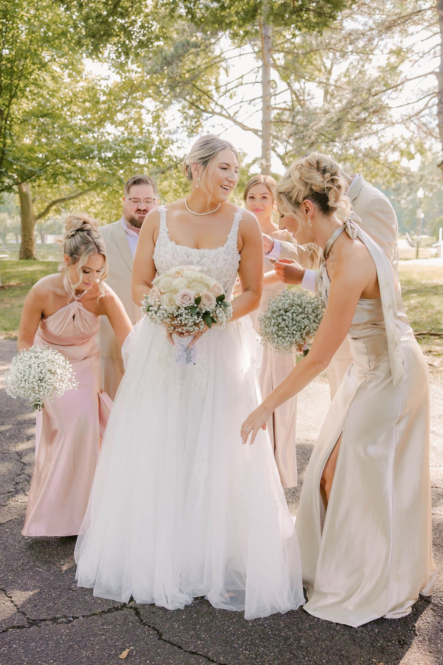 Bride in white gown surrounded by bridesmaids in blush dresses adjusting her skirt outdoors.