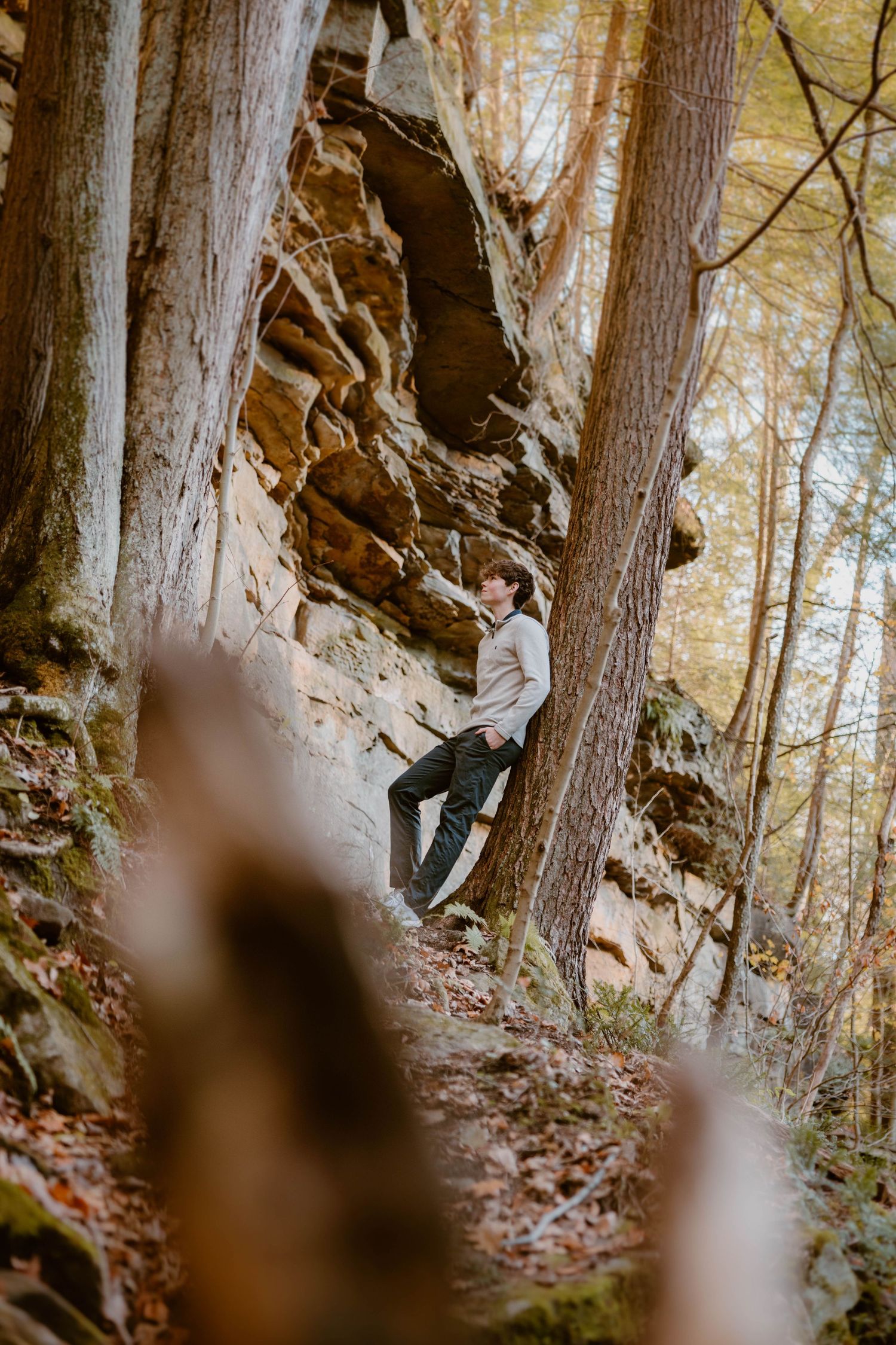Teen boy leaning against a large rock formation surrounded by tall trees in an autumn forest.