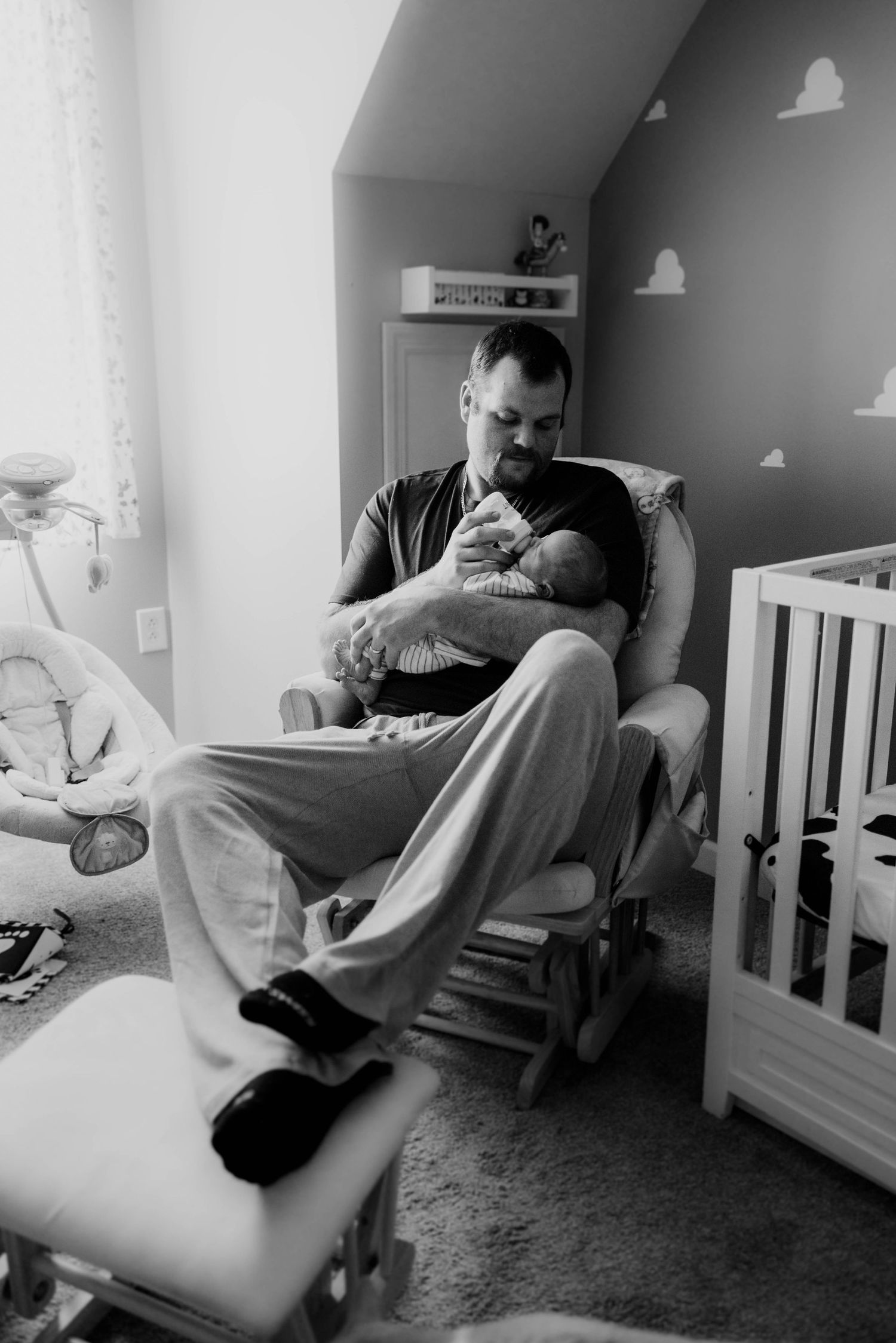 Black and white photo of a father sitting in a rocking chair holding his newborn in a nursery.