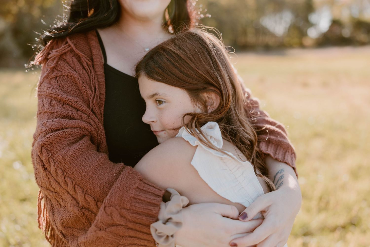 A mother in a rust cardigan tenderly embraces her young daughter in warm golden sunlight.