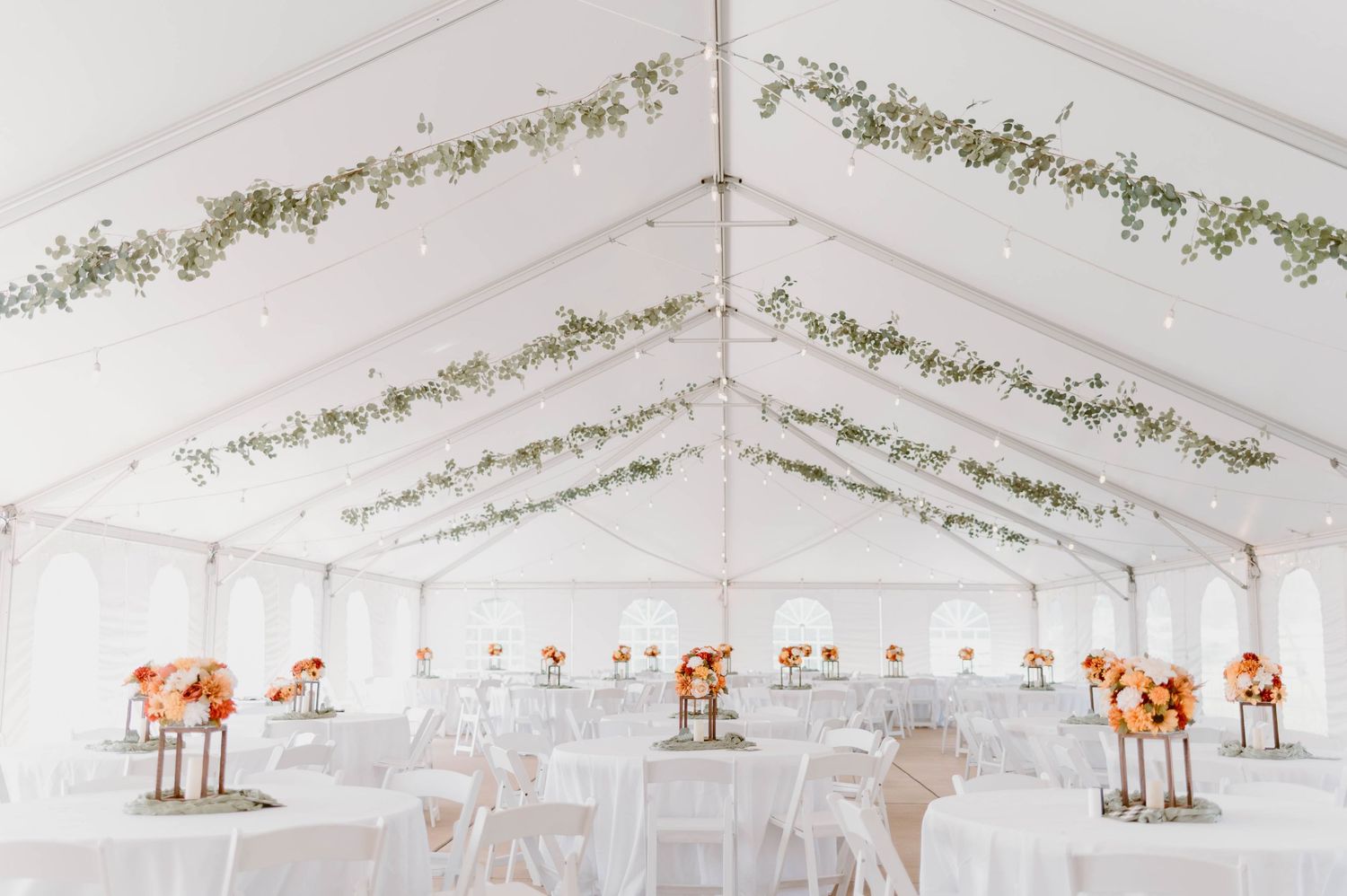 Elegant wedding reception tent with white tables, orange floral centerpieces, and greenery garland ceiling decor.