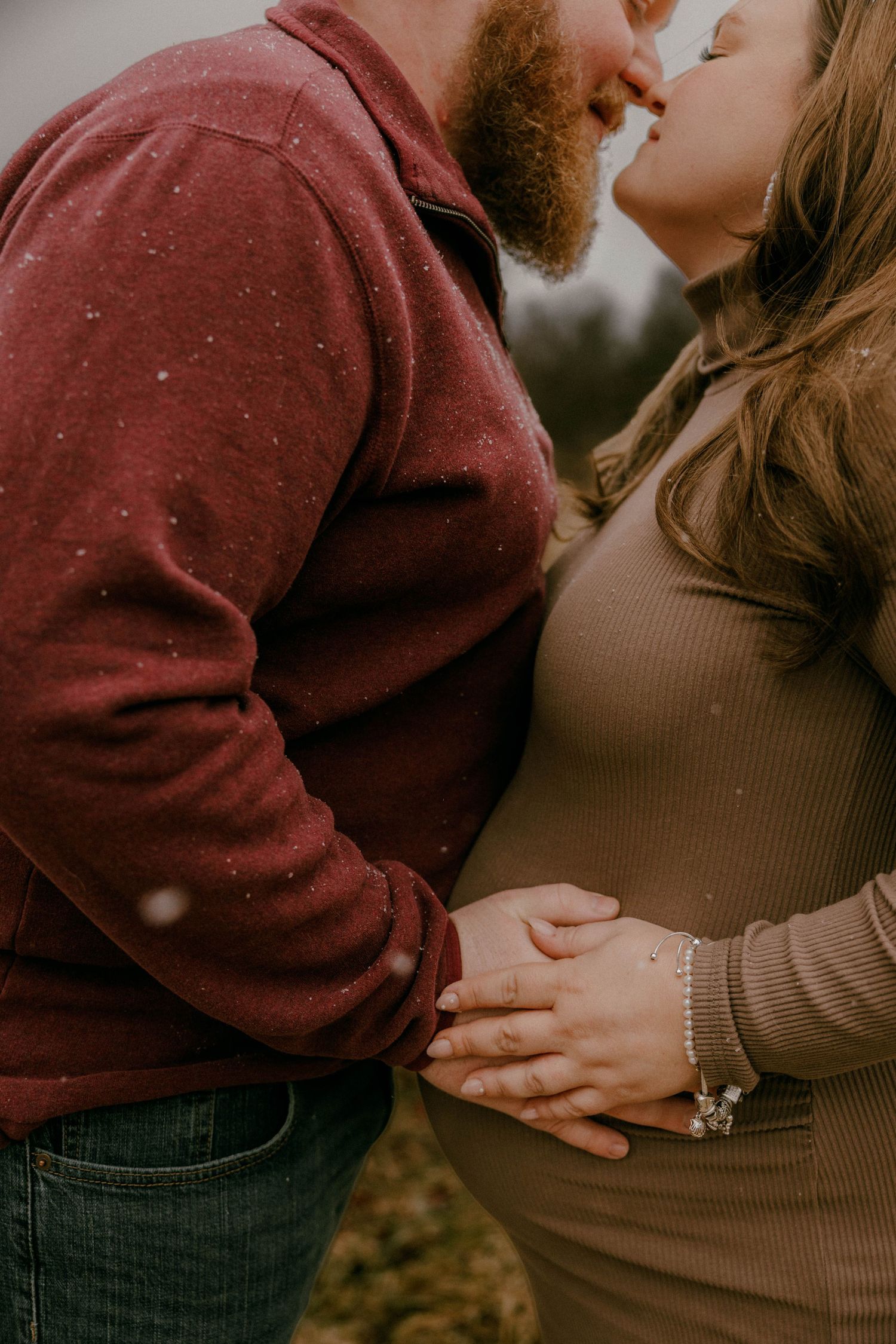 Parents smiling lovingly at their baby on a white blanket in a green meadow.