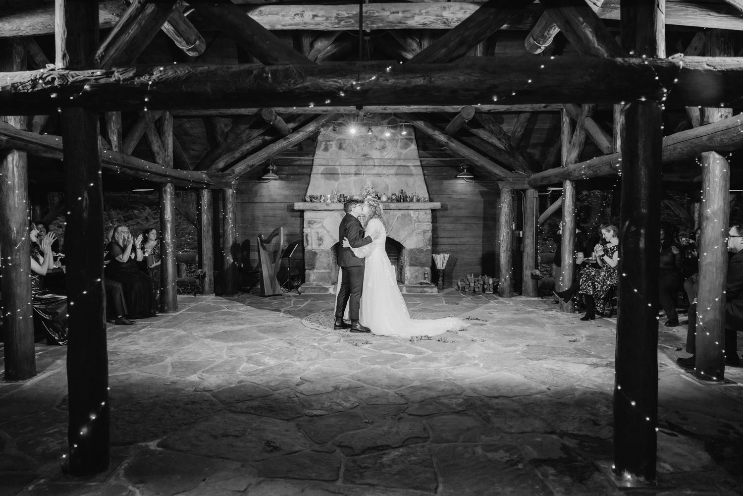 Black and white photo of bride and groom sharing first dance under rustic wooden pavilion with string lights and stone fireplace.