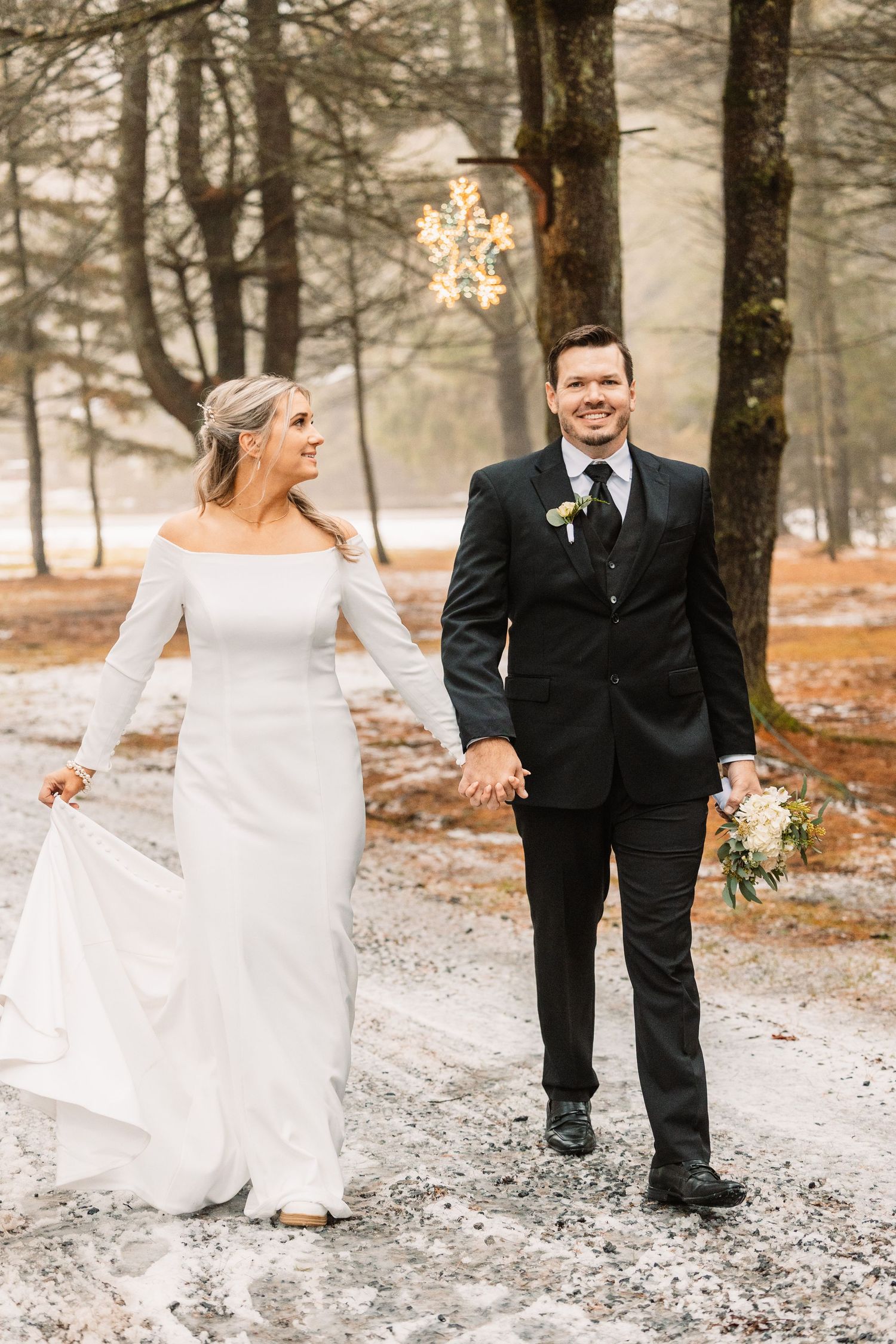 Smiling bride and groom walking hand-in-hand through snowy wooded path in winter wedding attire.