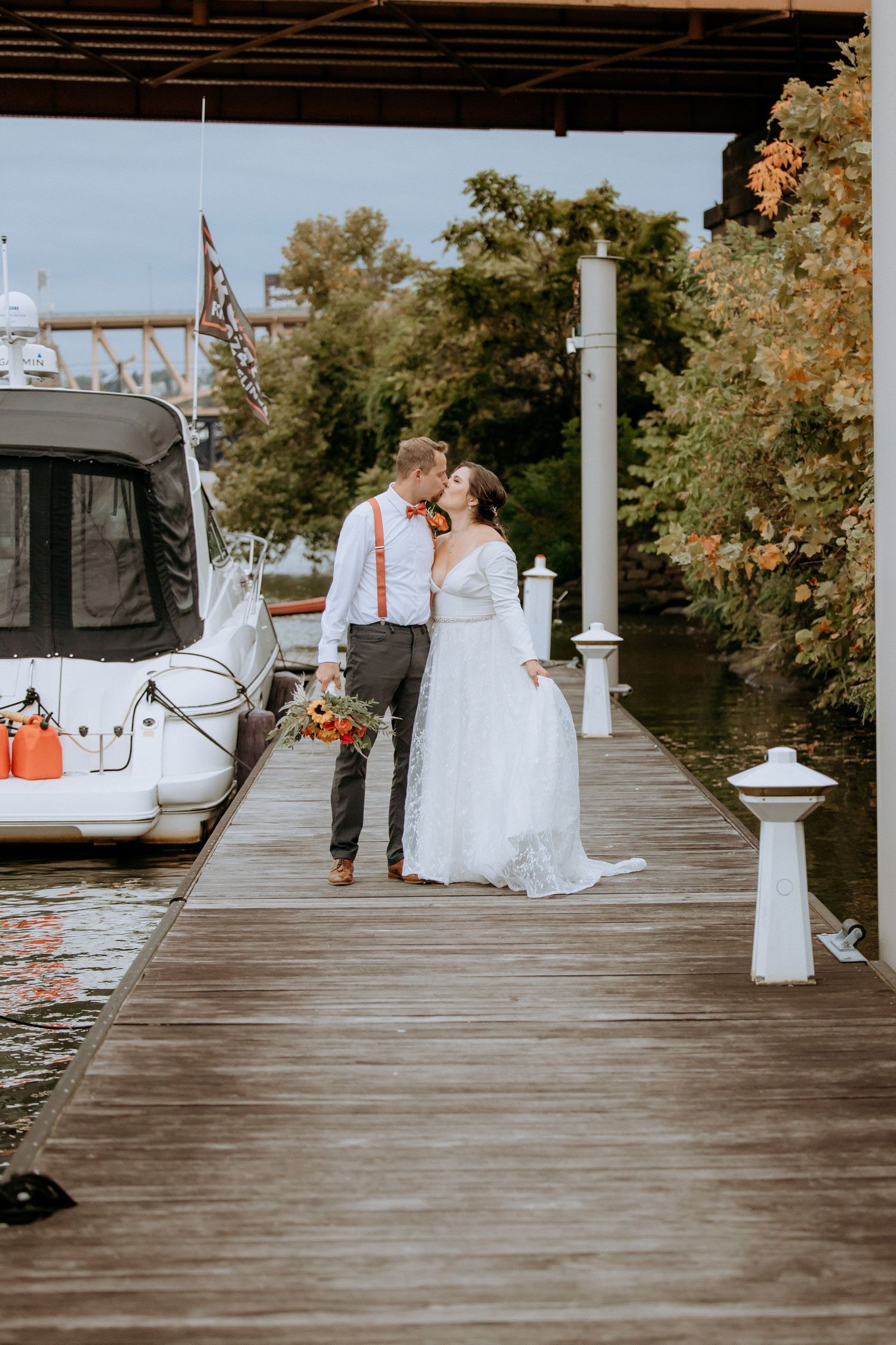 Bride and groom kissing on a wooden dock beside a yacht with autumn trees and calm water in background.