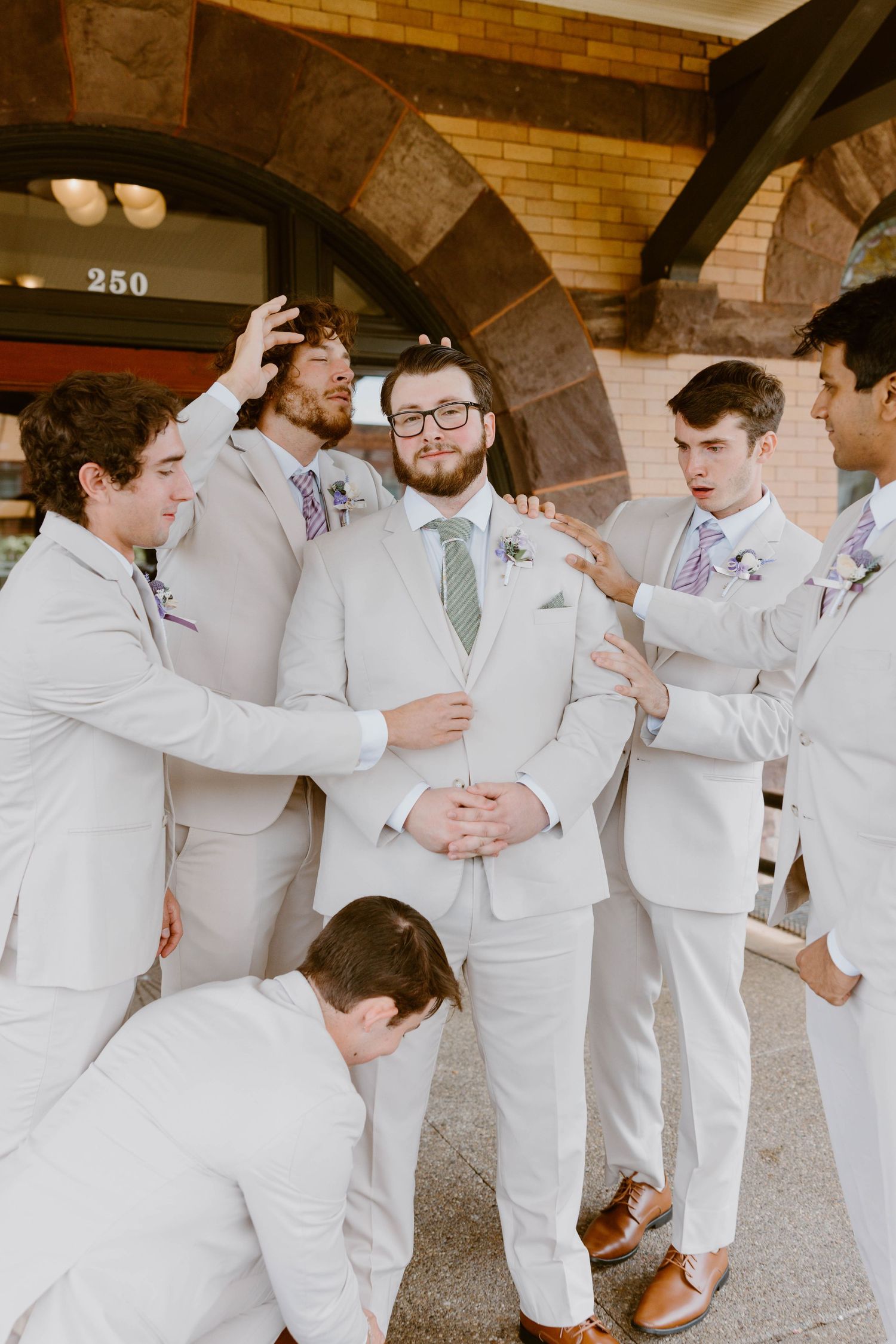 Groom in light gray suit surrounded by groomsmen in matching suits outside a brick building entrance.