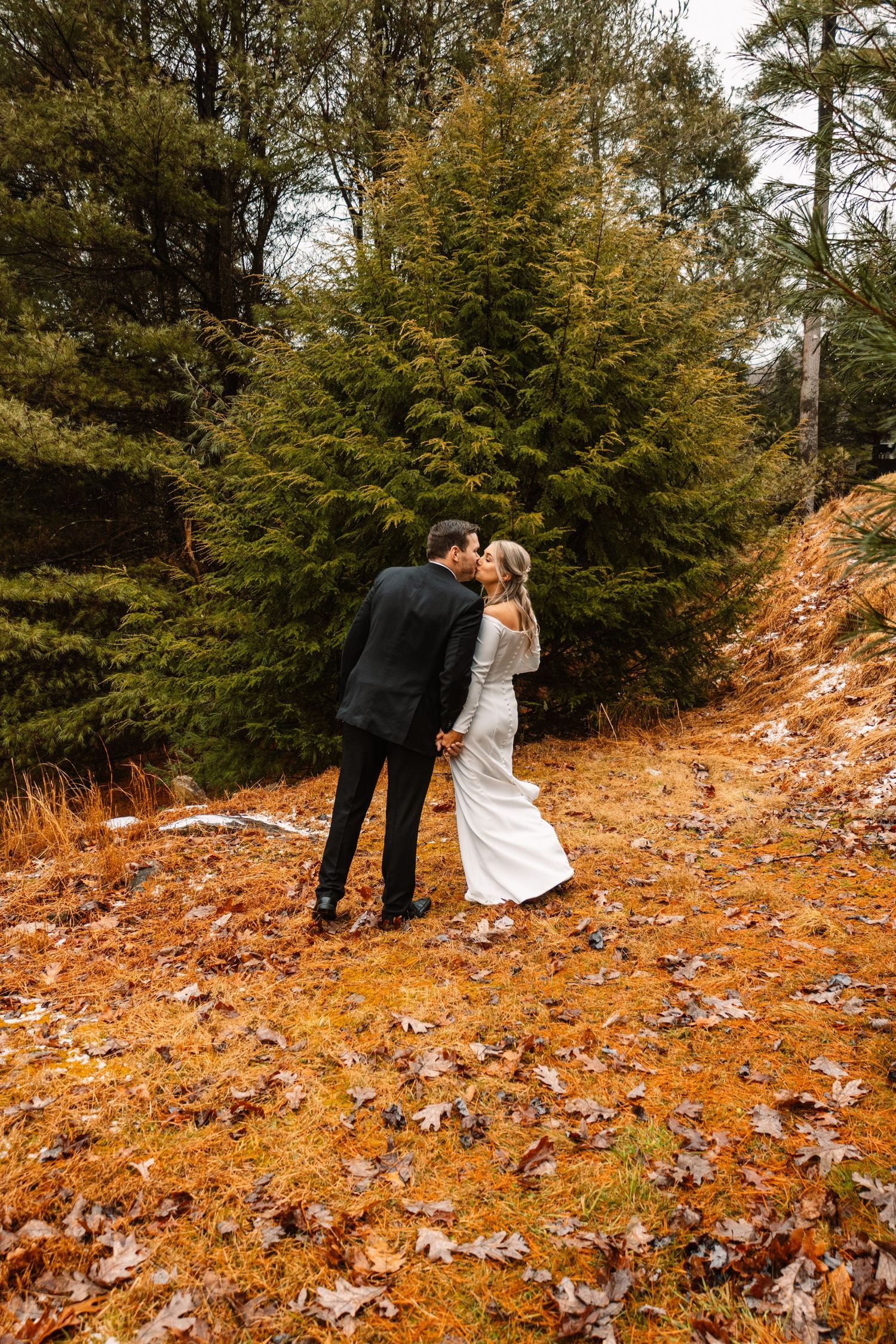 Bride and groom share a romantic kiss on a forest hillside covered in autumn leaves during their wedding day.