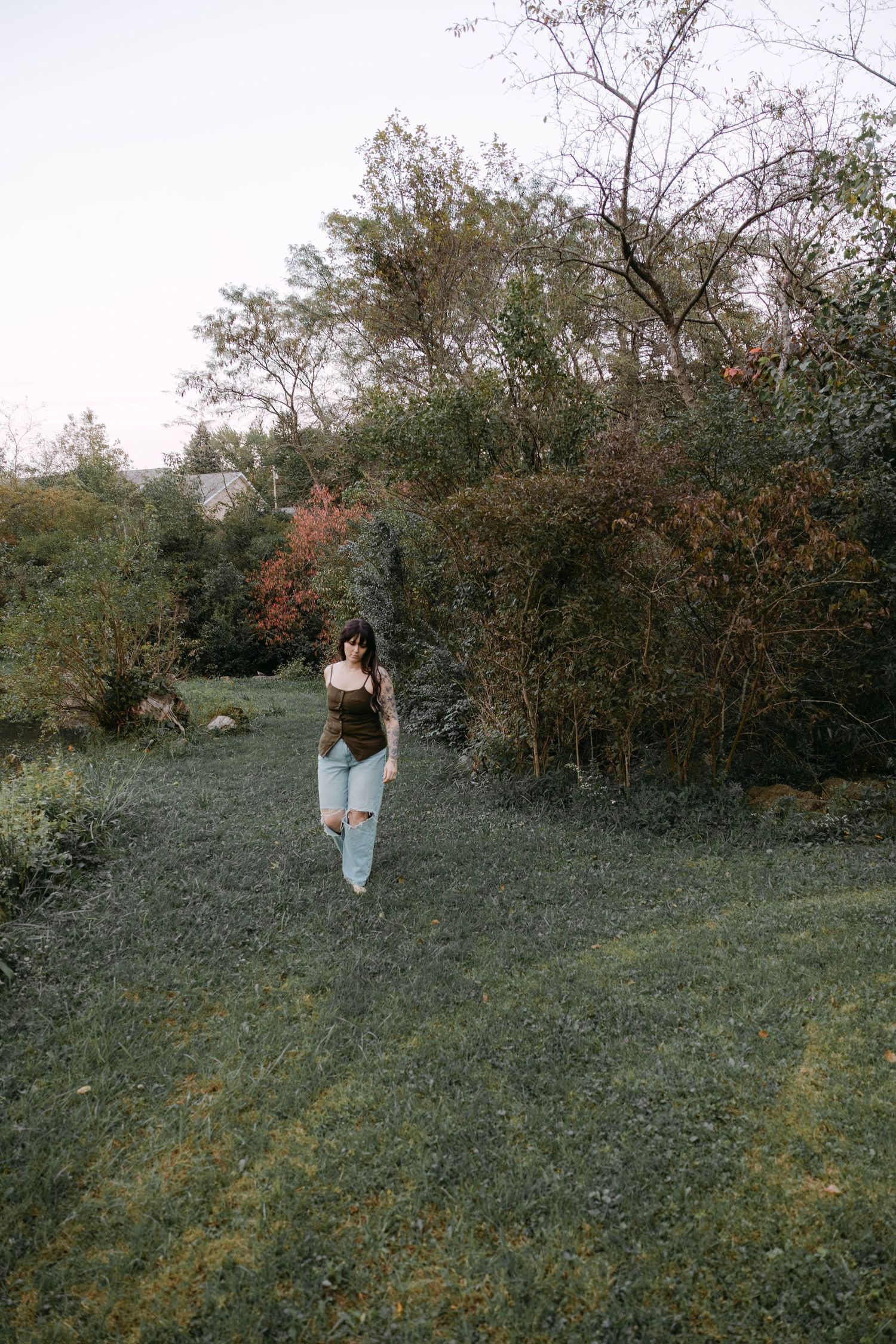 Woman in a green top and ripped light-wash jeans walks barefoot across a grassy lawn lined with autumn shrubs and trees.