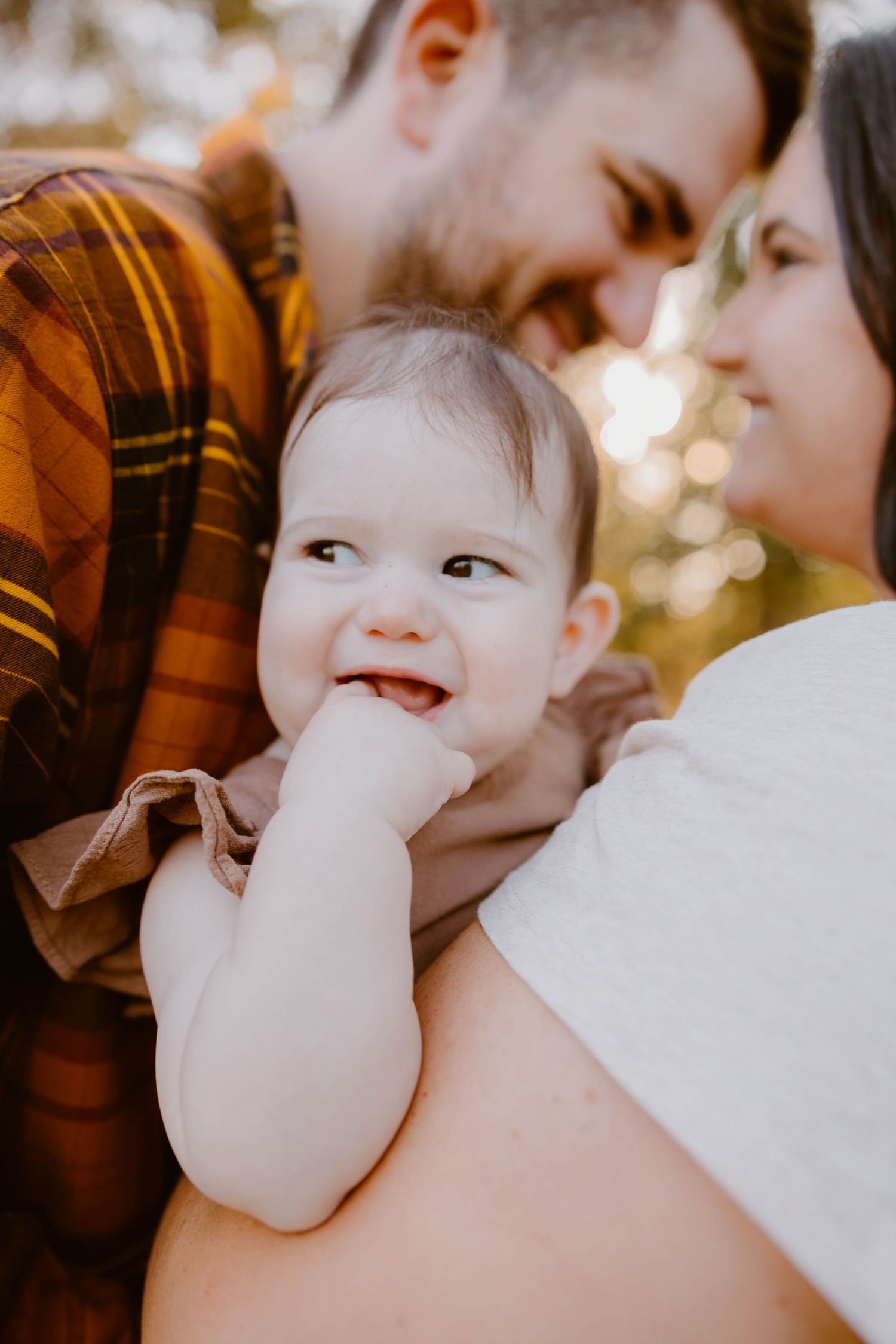 Smiling baby looks up while parents lean in closely during a warm outdoor family portrait.
