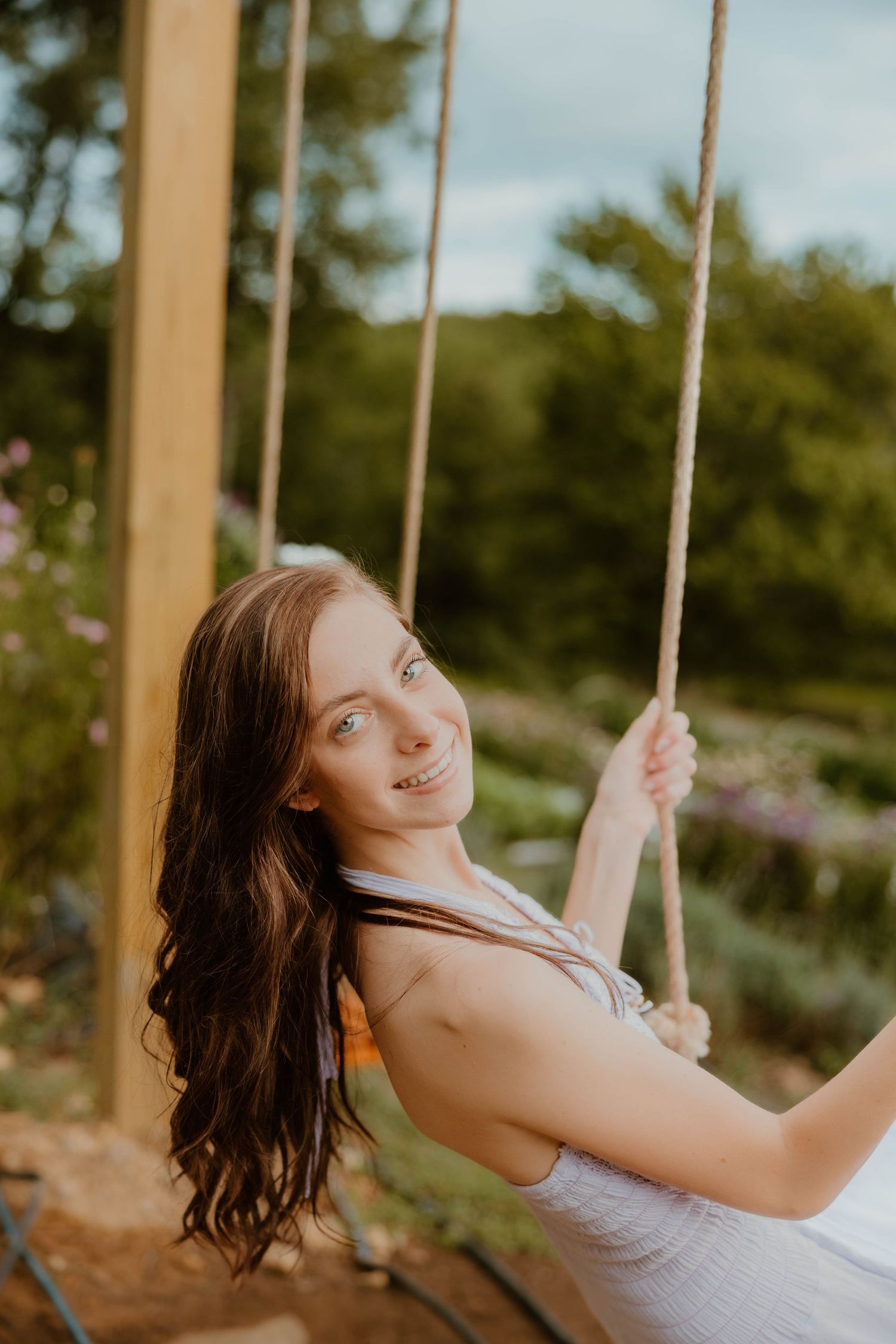 Smiling young woman with long brown hair sitting on a rope swing in a sunny outdoor garden setting.