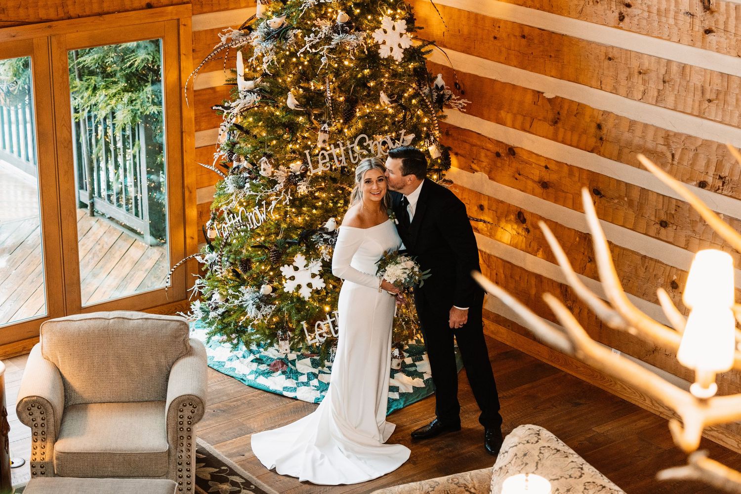Bride and groom pose together in a rustic log cabin beside a decorated Christmas tree on their winter wedding day.