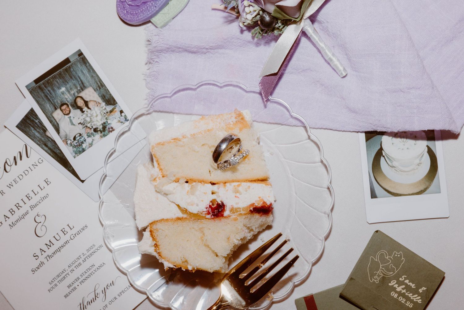 Wedding flat lay with cake slice, rings, invitation, polaroid photos, and purple napkin on white surface.