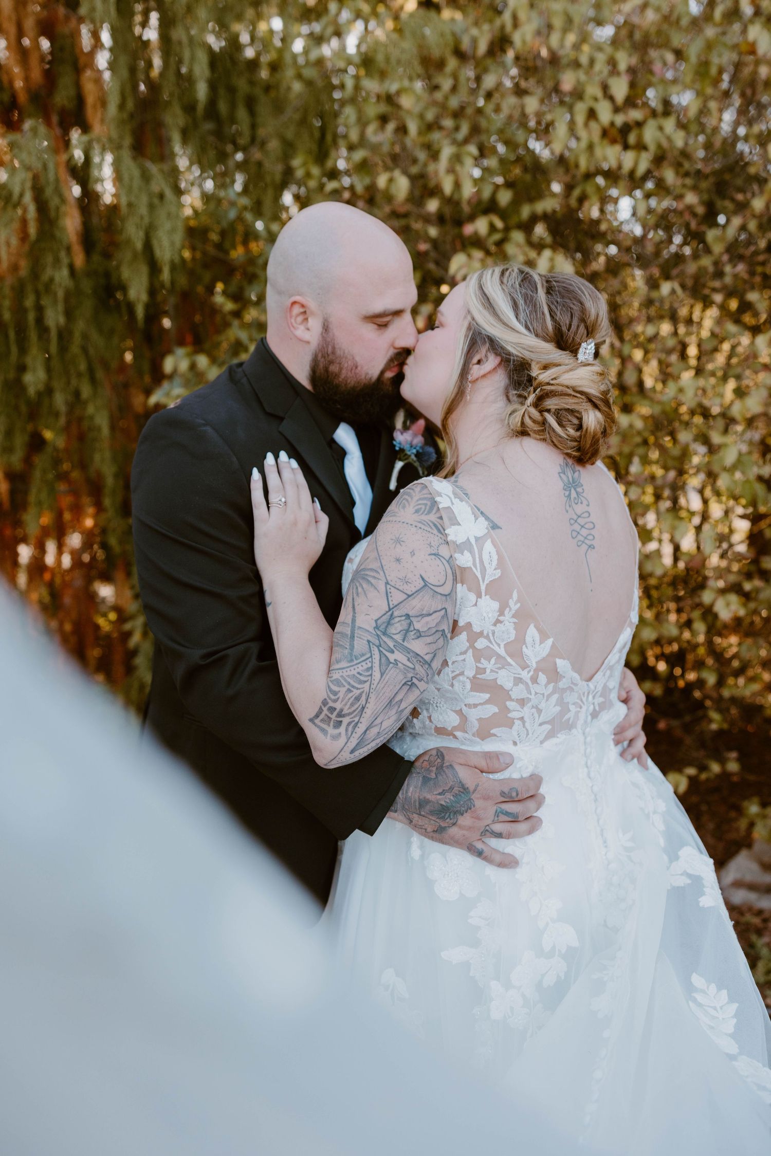 Bride and groom share a kiss outdoors, bride wearing a lace backless gown with sleeve tattoos visible at the Atrium in Butler, Pennsylvania.