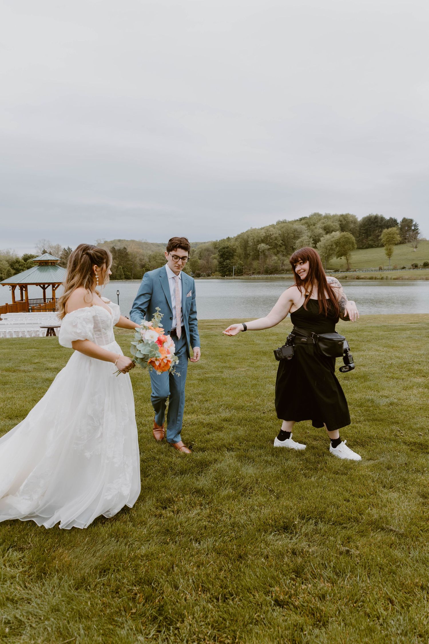 Photographer directing bride in white gown and groom in blue suit during outdoor wedding near a scenic lake.