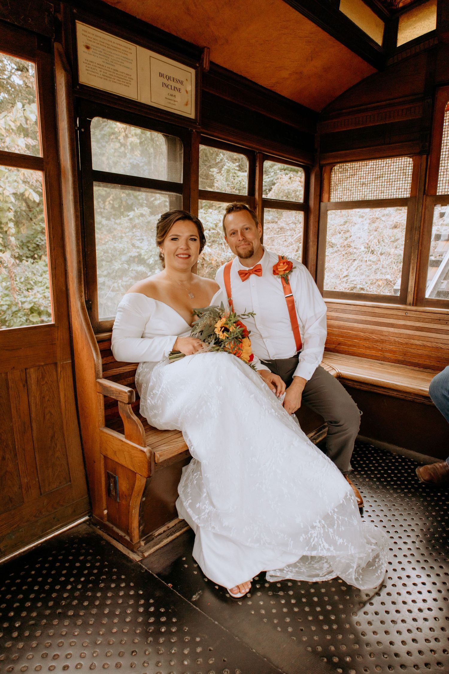 Bride and groom sit together inside a vintage wooden trolley car, bride holding a sunflower bouquet.