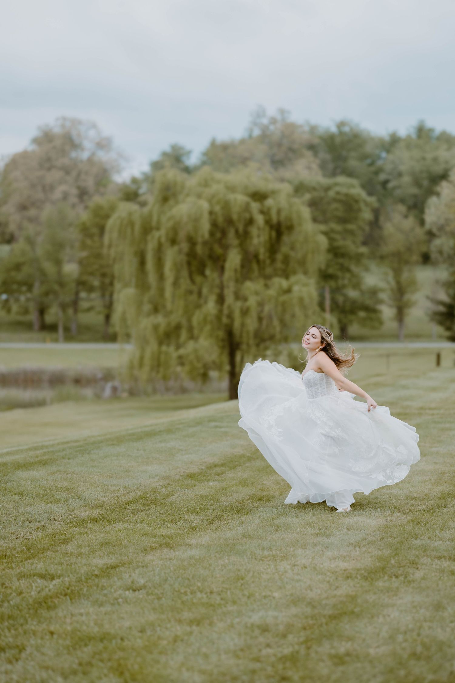 Bride twirls in a flowing white tulle wedding gown on a green lawn with willow trees in the background.
