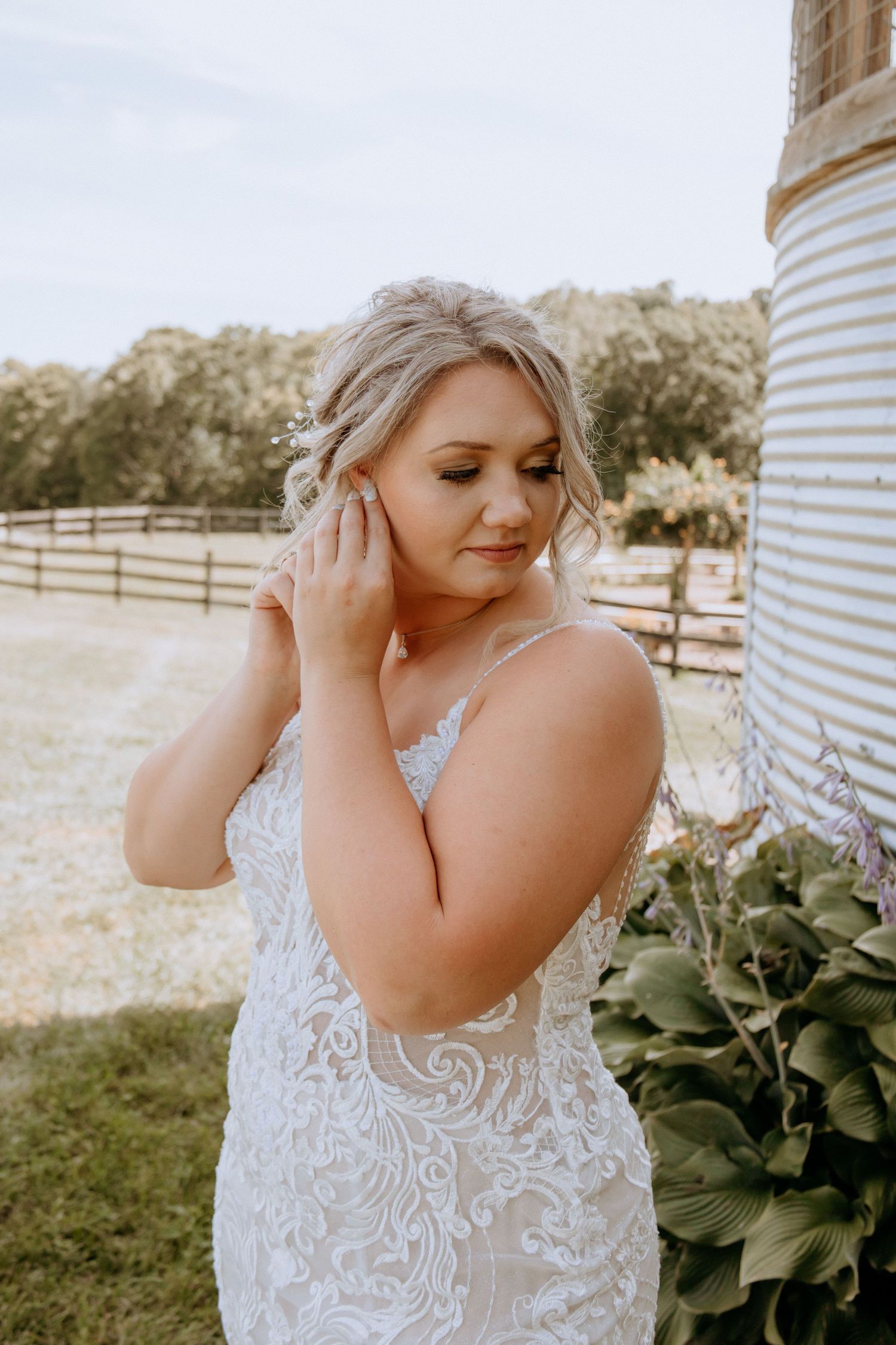 Blonde bride in embroidered lace gown adjusts earring beside a grain silo at a farm wedding venue.