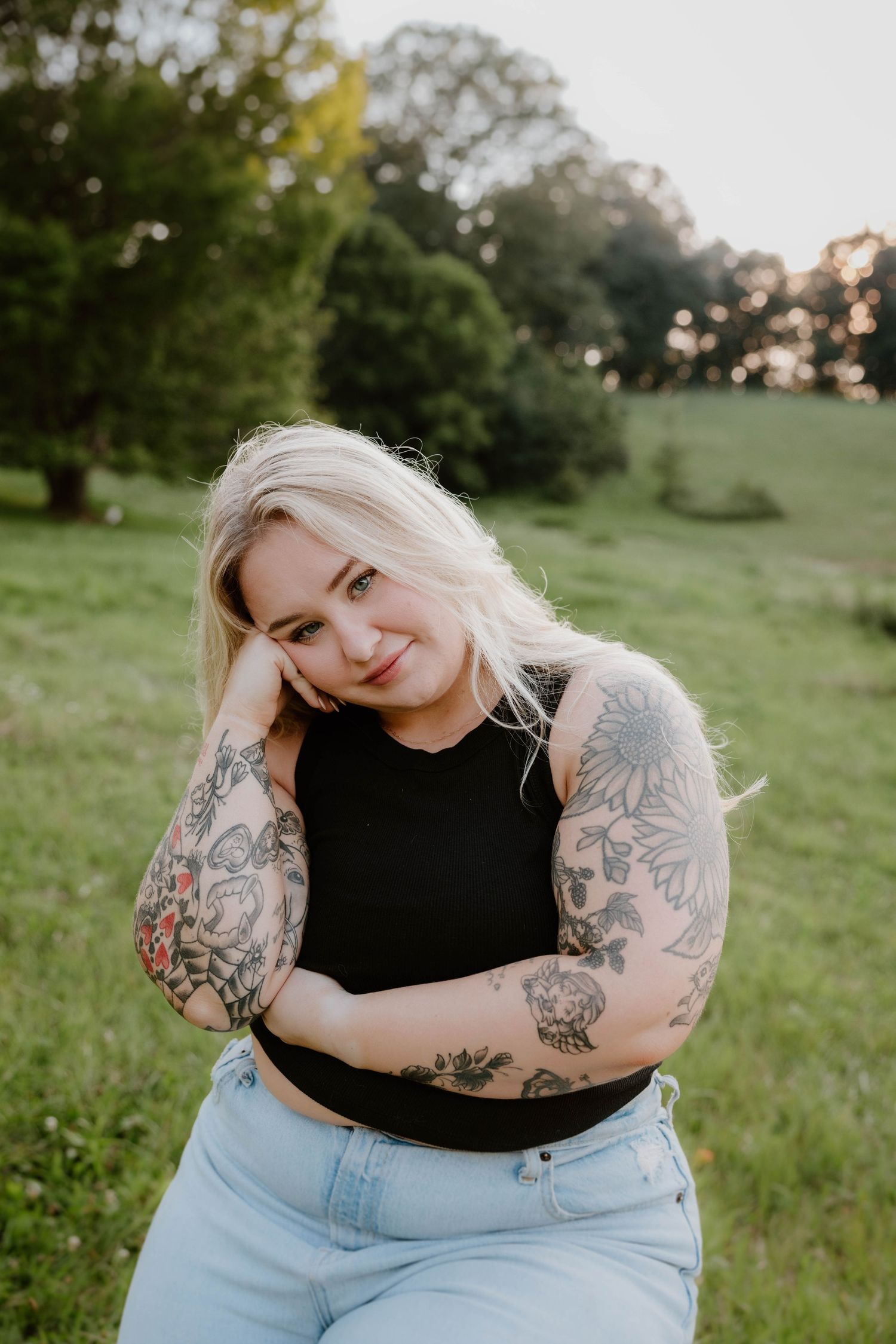 Blonde woman with floral tattoos on her arms posing in a green field during golden hour.