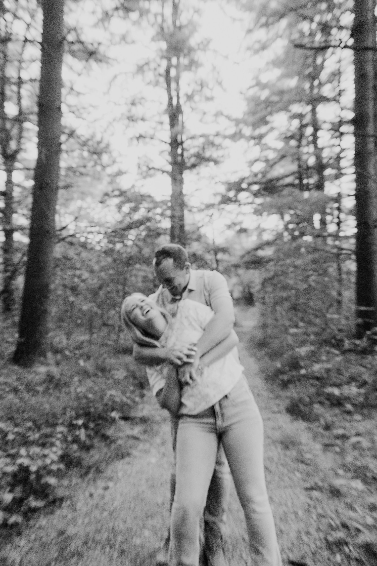 Black and white photo of a couple embracing and laughing on a wooded forest trail surrounded by tall pine trees.