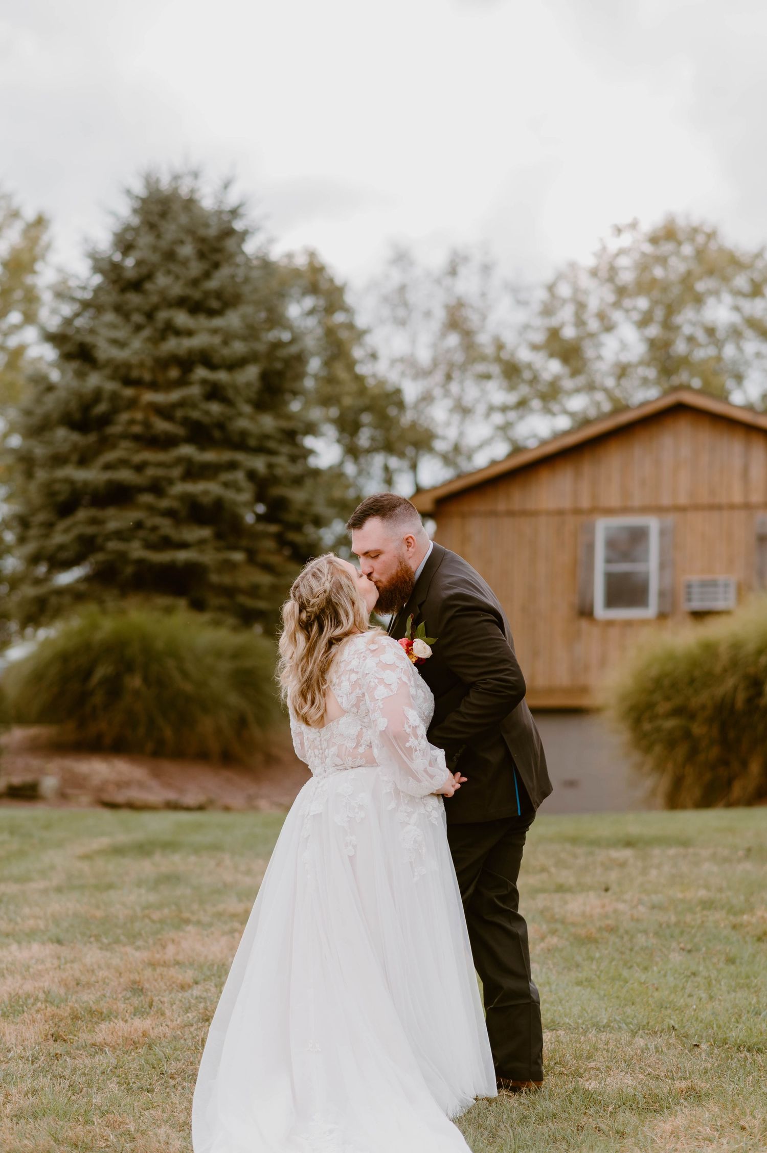 Bride and groom sharing a romantic kiss on green lawn with rustic barn backdrop.