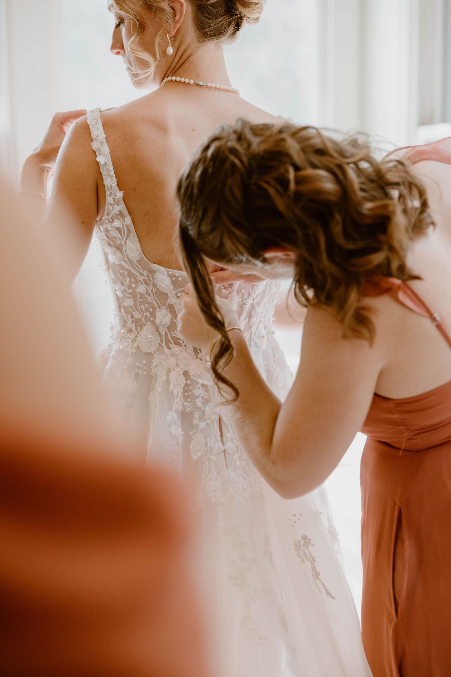 Bridesmaid in terracotta dress fastening the back of a lace bridal gown.