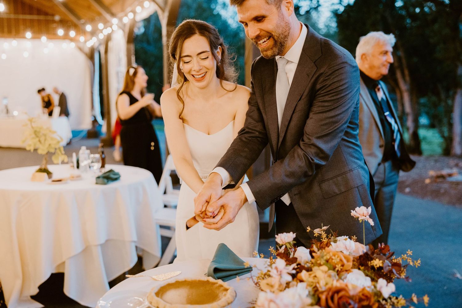Smiling bride and groom cutting wedding cake together at outdoor evening reception with string lights.