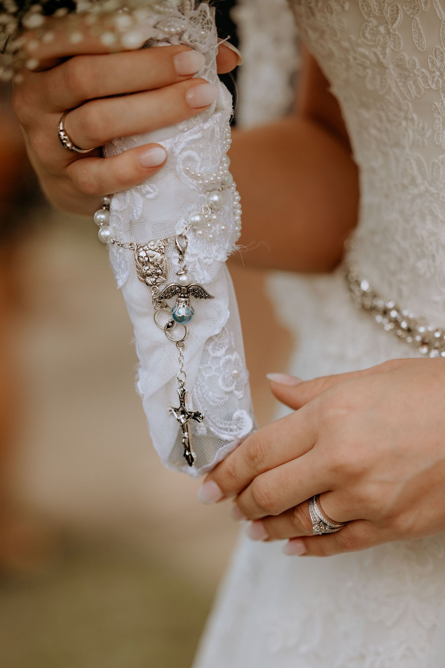 Bride holding decorative white lace garter with pearl and cross charms in close-up detail shot.